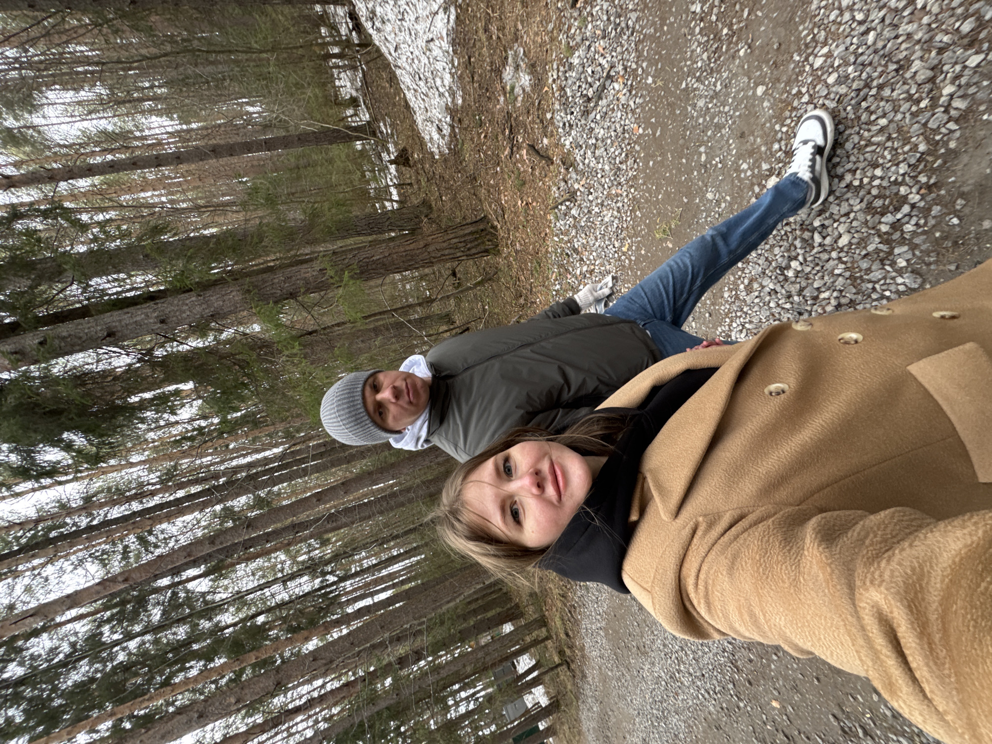 The image, taken at a tilted angle, shows two individuals on a path in a wooded area, likely a forest. In the immediate foreground, a woman with blonde hair and light-colored eyes looks towards the camera with a slight smile. She is wearing a black top or hoodie underneath a warm, light brown (camel-colored) coat with visible buttons.
Behind her and slightly to the left, a man is visible. He is wearing a grey knit beanie, a white hoodie, and a dark grey or olive green puffer jacket. He has blue jeans and white and black sneakers on his feet.
The setting appears to be a natural environment with numerous tall, slender trees, possibly pines or similar evergreens, dominating the background. Their trunks are visible, and some have sparse green foliage. The ground they are walking on is a mix of dirt and gravel, with patches of melting snow and dried leaves or brush. The presence of snow suggests it is late winter or early spring. The lighting is soft and natural, indicating daytime, possibly an overcast day. Both individuals seem to be enjoying a casual outdoor walk. There is no visible text in the image.