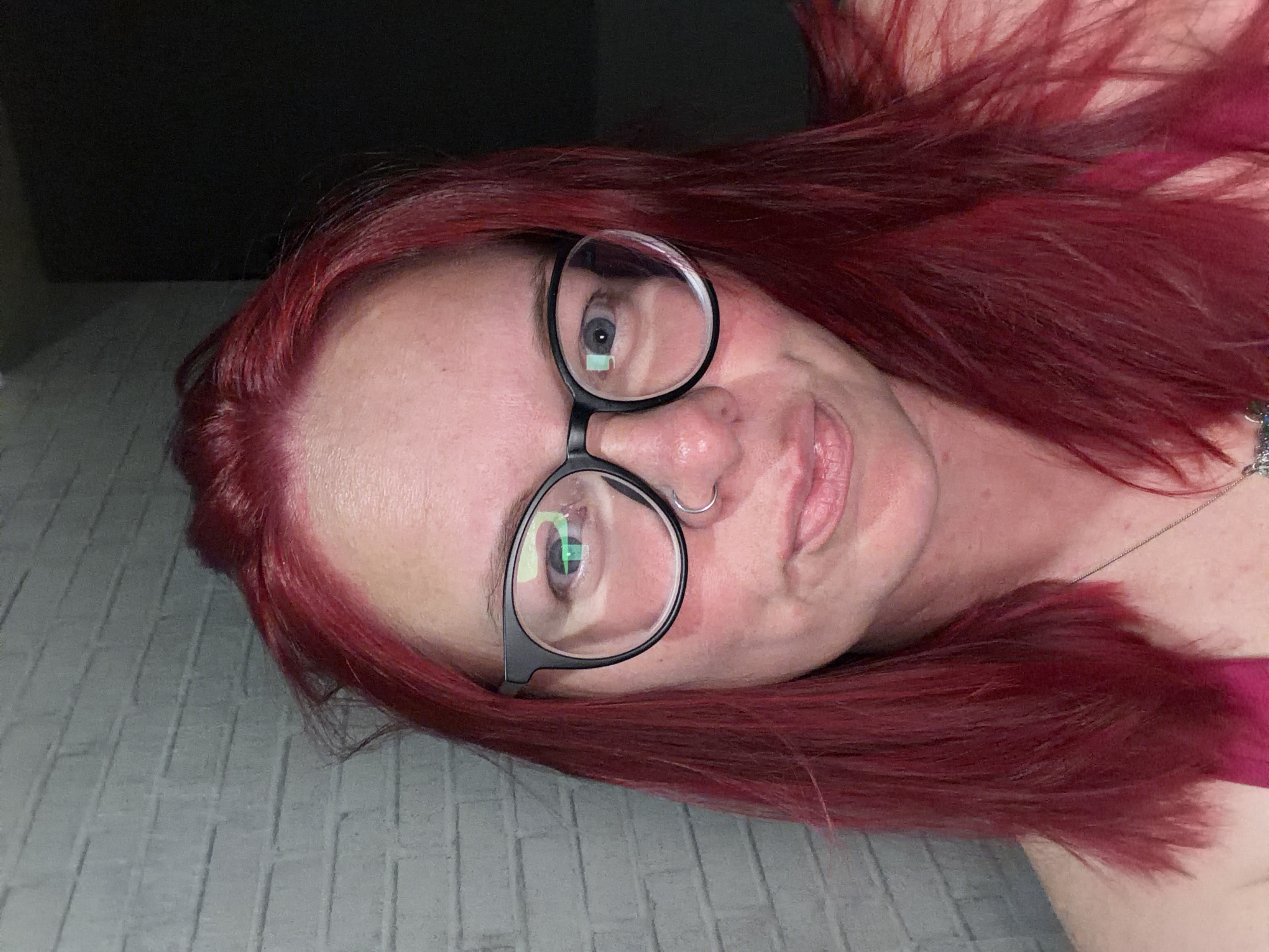 A close-up, sideways portrait features an individual with fair skin and vibrant red hair, lying on their right side, looking directly at the camera. The person is wearing black-rimmed eyeglasses, which reflect light sources, including a distinct green reflection in the left lens over the eye. A silver hoop nose ring is visible on their left nostril, and a delicate silver chain necklace is around their neck. They wear a magenta or dark pink garment, partially visible on the right side of the frame. The individual has a slight smile or smirk, conveying a relaxed and neutral expression. Their red hair is fanned out around their head. The background consists of a light gray, textured surface that appears to be a bedspread or upholstered furniture, while the upper left portion of the image is a dark, indistinct area. No visible text is present.