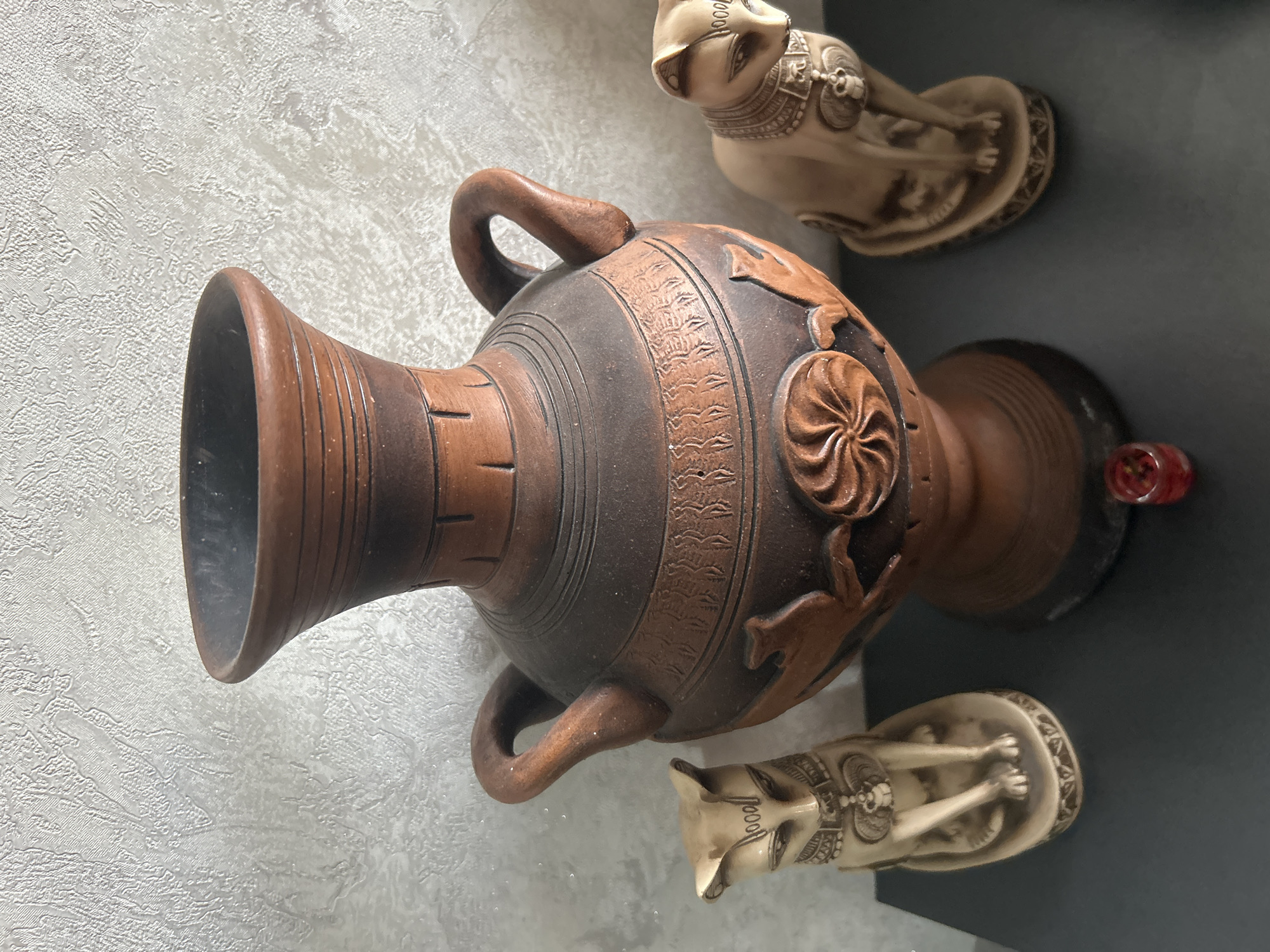 The image shows a collection of artifacts arranged in an indoor setting. An ornate, brownish-red vase with two handles is the central object. The vase has darker brown or black accents around the neck and base, with decorative carvings on the body. Two small, beige Egyptian cat figurines are positioned near the vase, one above and one below. The setting appears to be against a wall on the left, and a gray surface on the right. Without further context, it is impossible to determine its specific location in Tbilisi, Georgia.
