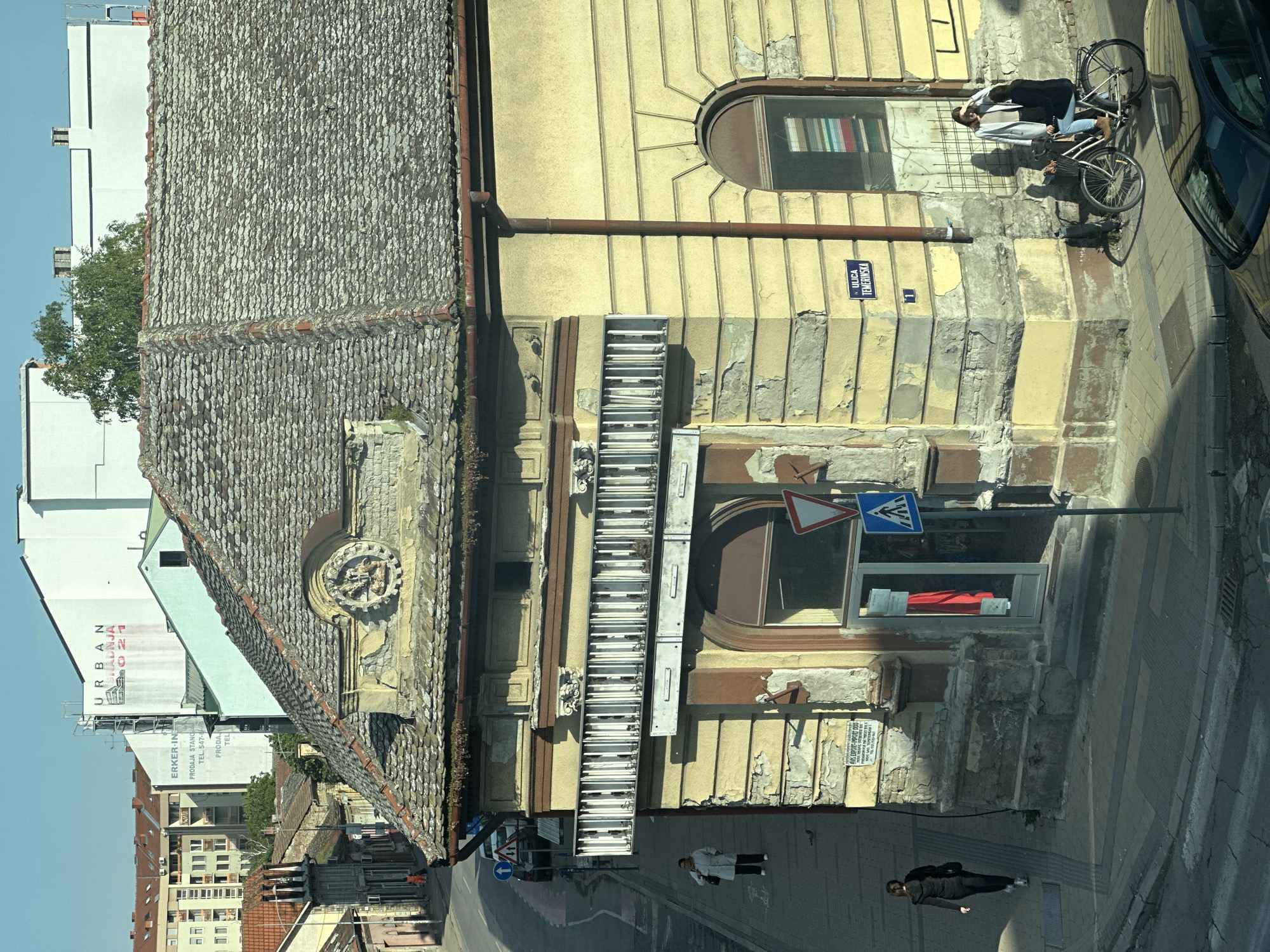 The image displays a sunlit street scene in Novi Sad, Serbia, characterized by a mix of historical and contemporary architecture. On the right, a prominent old yellow building with significant peeling paint features a grey, textured tiled roof and a decorative relief on its gable. A brown downpipe runs along its side. Behind this structure, modern white buildings are visible, one prominently displaying "URBAN GRADNJA 021" and another with "ERKER-IN PRODAJA STANOV..." text and a phone number (TEL. 547-). The clear blue sky and sharp shadows suggest a sunny mid-day.
Everyday urban activity is captured on the sidewalk. Three individuals are visible: one person stands by a bicycle, wearing a light jacket and dark trousers. Nearby, a blue street sign clearly reads "ULICA TEMERINSKA 1". Further down the street, another person in dark clothing walks away from the camera, and a third individual in a light coat stands closer to the foreground.
Various objects and signs populate the scene. A long, rectangular fixture with multiple fluorescent tubes is attached to the yellow building's facade. A round window on an upper floor of the yellow building reveals a colorful arrangement that appears to be book spines. Additional street signs include a pedestrian crossing sign, a red triangular yield sign, and a partially visible blue 'turn right' sign. A brown car is partly visible on the far right. A sign on the yellow building also reads "AMS EXPORT-IMPORT DOO". The scene offers a candid view of urban life, highlighting architectural contrasts and routine activities.