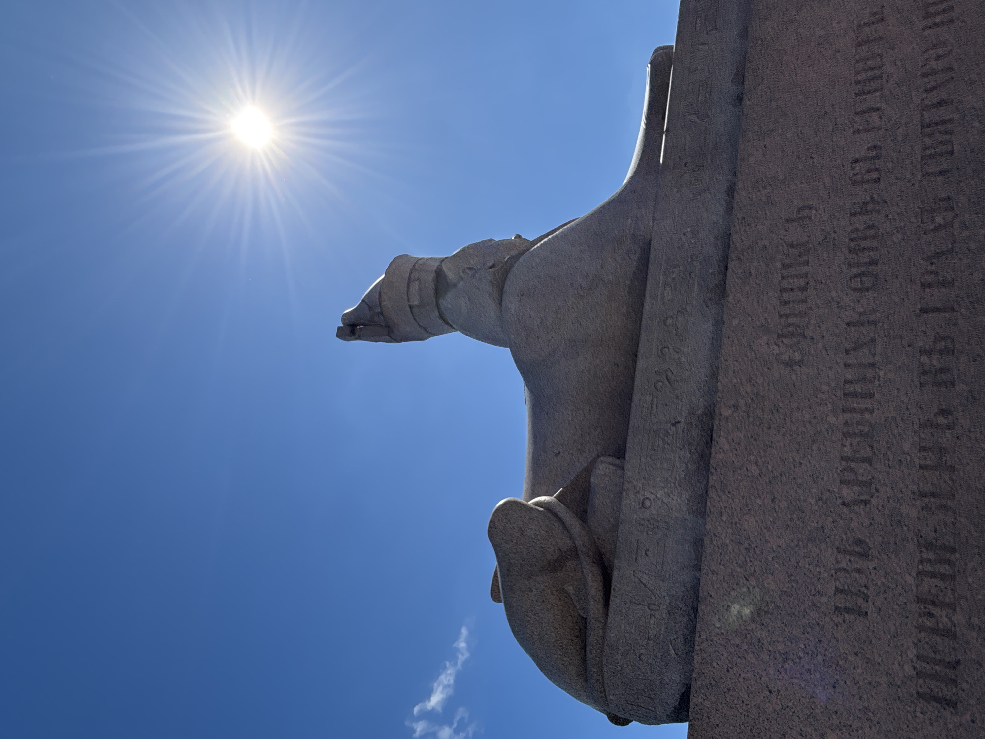 The image captures a stone sphinx statue against a bright blue sky with the sun prominently visible and radiating light. The sphinx is seen from a low angle, emphasizing its scale and detail, with hieroglyphs etched onto its base. The inscription on the granite base is in Cyrillic script. The statue is likely located in Saint Petersburg, Russia, potentially on the University Embankment or the Imperial Academy of Arts, where similar sphinxes are displayed. The environment appears to be outdoors on a clear, sunny day.
