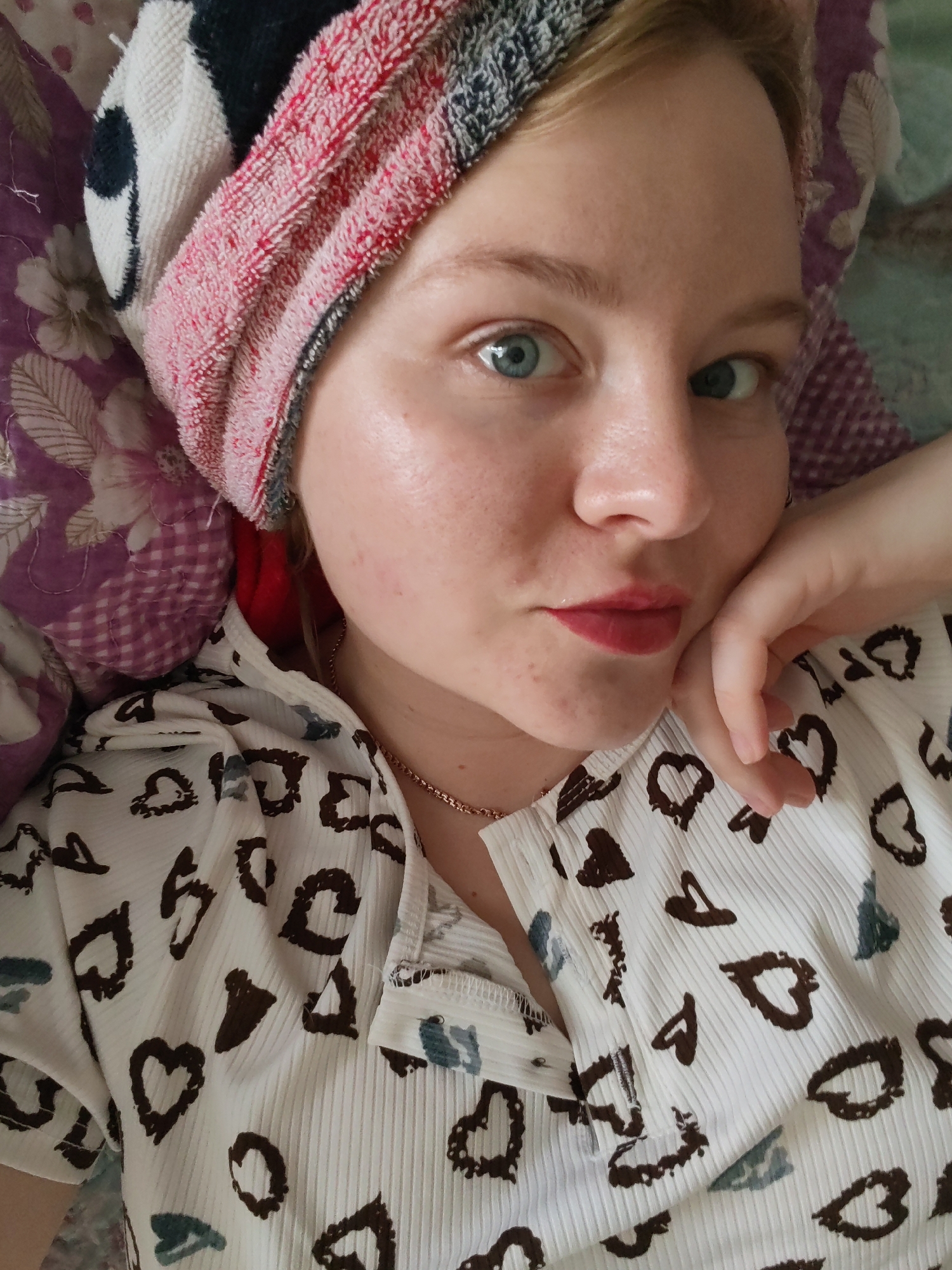 A young, fair-skinned woman is lying down, propping herself up with her right hand under her chin. She has light blue eyes and is wearing red lipstick. A multi-colored towel is wrapped around her head. She is wearing a white shirt patterned with brown hearts and blue accents. A gold necklace is visible around her neck. She appears to be resting on a purple pillow with a floral pattern. The image does not contain information to verify if the location is in Izhevsk, Russia.