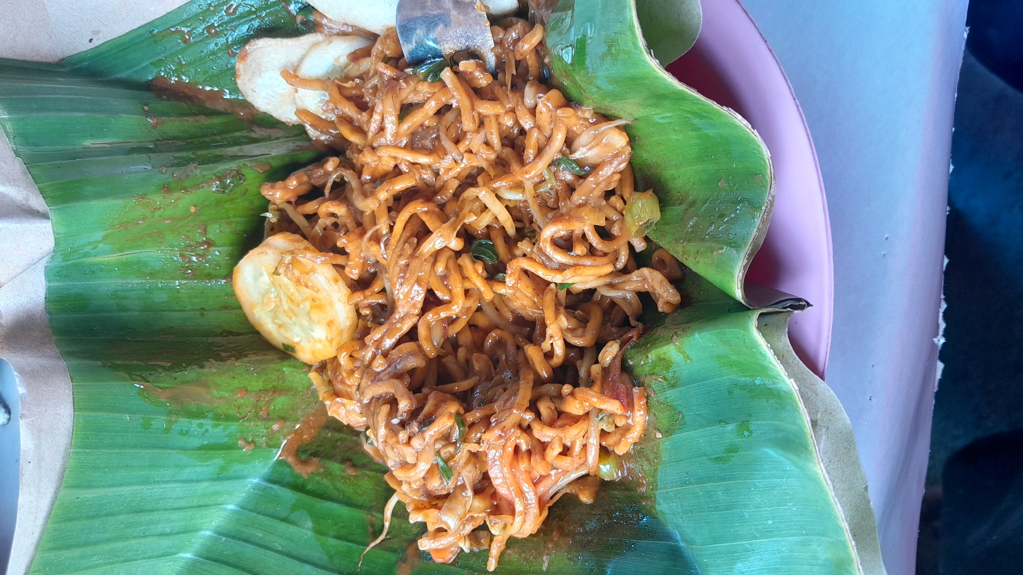 A top-down, close-up shot shows a portion of Mie Aceh, a spicy noodle dish from Aceh, Indonesia, served in a banana leaf. The noodles are thick, yellow, and coated in a dark reddish-brown sauce. Visible within the noodles are green herbs and small pieces of vegetables. To the left of the noodles are half-slices of hard-boiled eggs and what appears to be sliced potato or similar vegetables. A metal fork is partially visible near the top of the frame, embedded in the noodles. The banana leaf is shiny, wet, and vibrant green. The dish is resting on a pink plate, a small portion of which is visible on the right side. In the background, a dark blue or gray surface is seen, likely the table or surrounding area. The overall presentation suggests a street food or casual dining setting in Lhokseumawe, Indonesia.