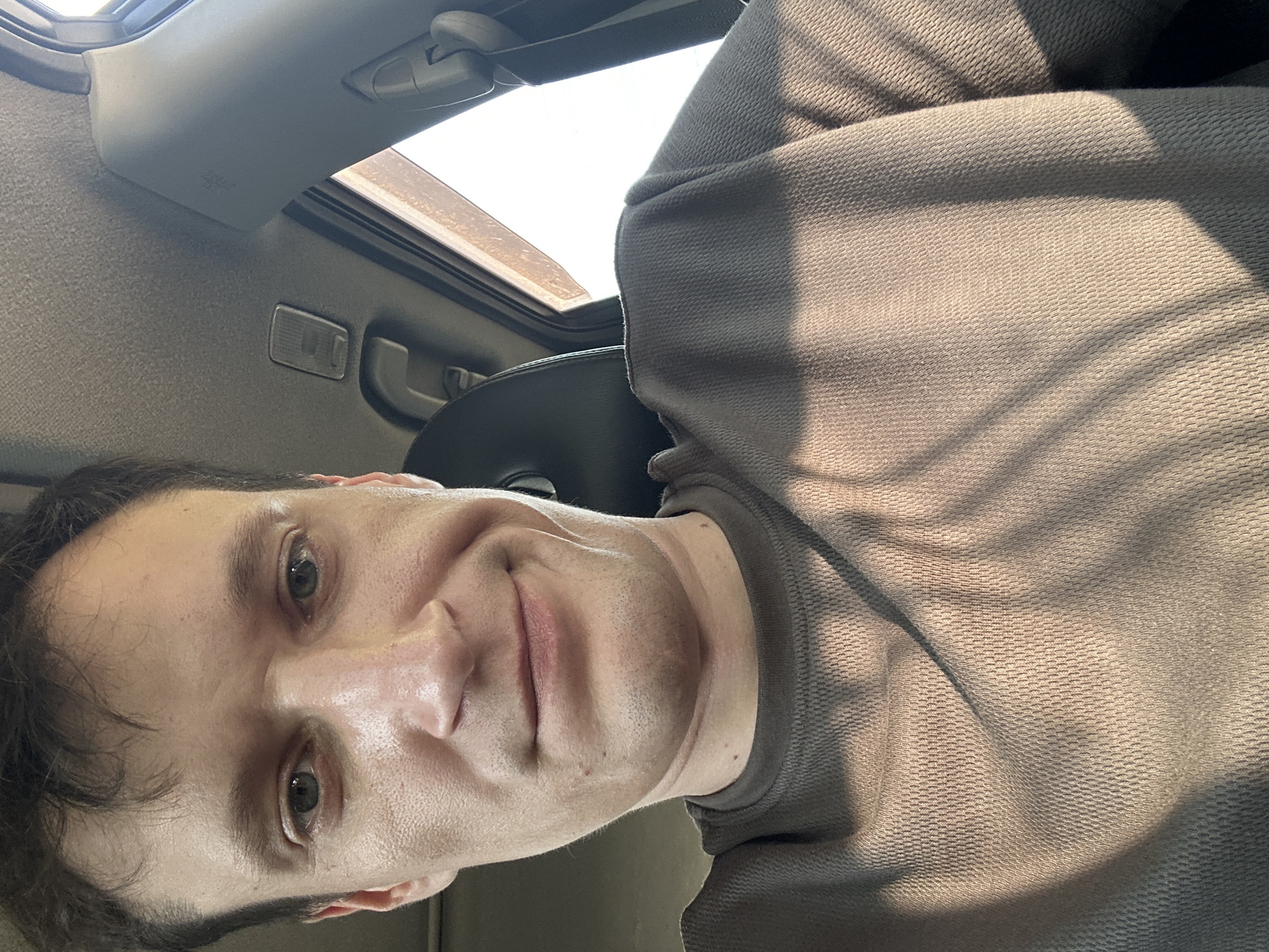 A close-up, selfie-style photo shows a man in the driver's seat of a vehicle. The man has fair skin, brown hair, and appears to be in his late 20s or early 30s. He is wearing a grey, long-sleeved t-shirt. He is smiling subtly. The interior of the car is visible, including a dark grey headrest and the car's ceiling. Sunlight streams in from the right side of the vehicle, casting shadows across the man's face and t-shirt. The man is positioned in the foreground, with the car's interior filling the background. The photo was taken in Makiv, Ukraine.