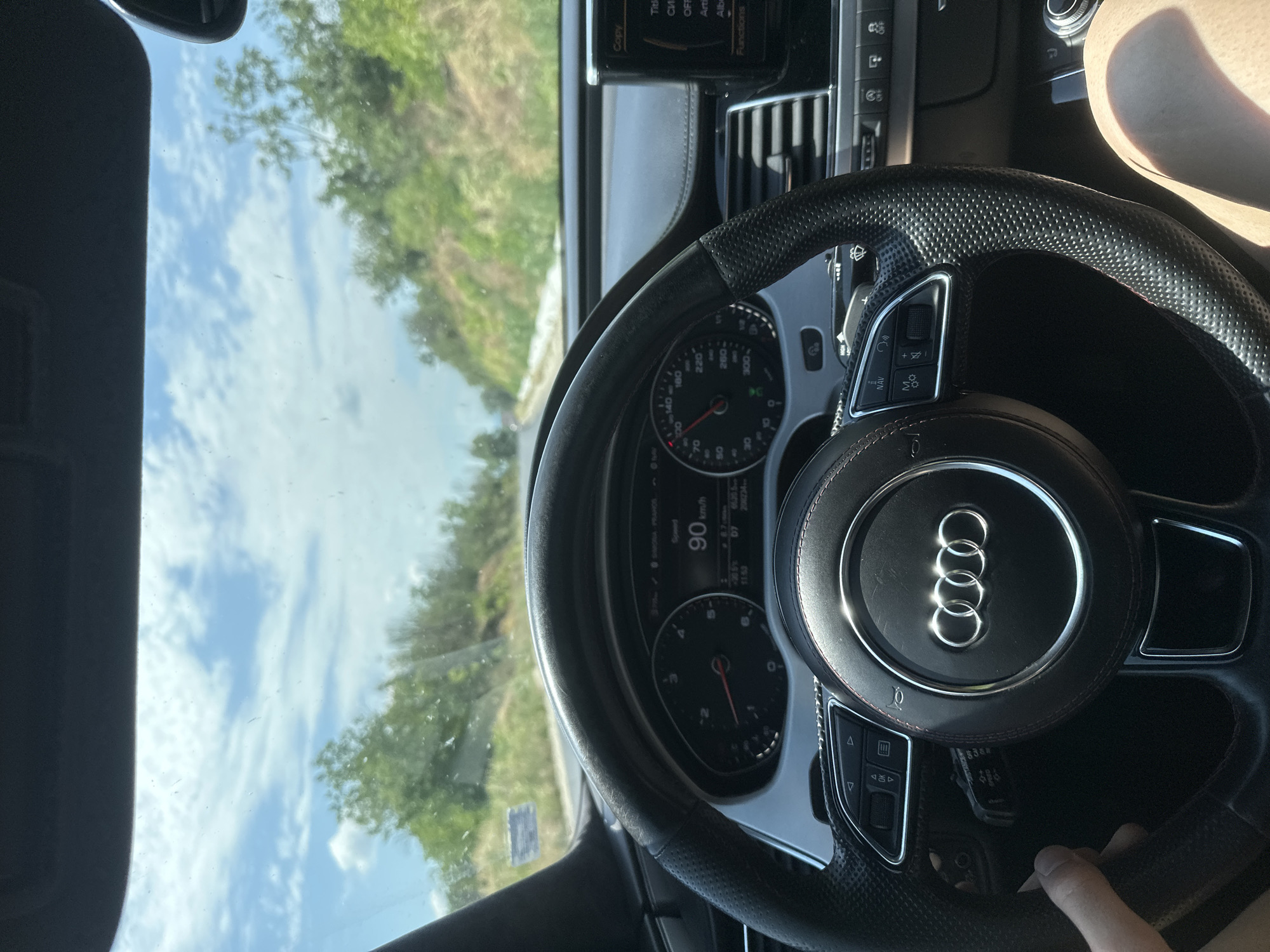 A first-person perspective shot from inside an Audi, shows the steering wheel and dashboard. The car is traveling on a road lined with trees and vegetation under a cloudy sky. The dashboard displays the speed at 90 km/h. The scene suggests the vehicle is on a road outside of the city, in the vicinity of Polikraishte, Bulgaria, possibly on a sunny day.