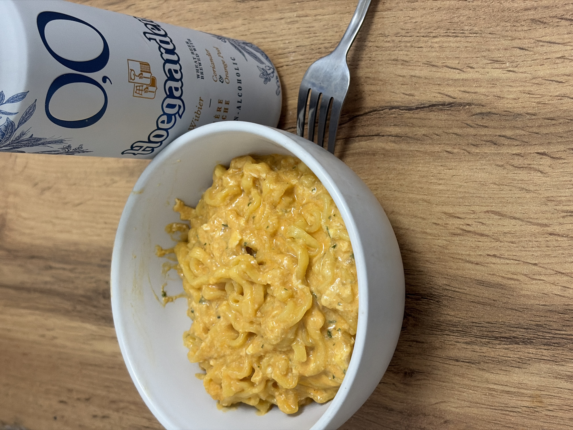 A top-down shot on a wooden surface shows a white bowl filled with macaroni and cheese, a fork, and a can of Hoegaarden 0.0 non-alcoholic beer. The beer can sits partially behind the bowl, tilted to the left, with its label facing upward. The fork is next to the bowl, with the tines angled towards the center. The macaroni and cheese appears creamy and slightly orange in color. No people are visible. The image does not contain information about Chaikovsky, Russia.