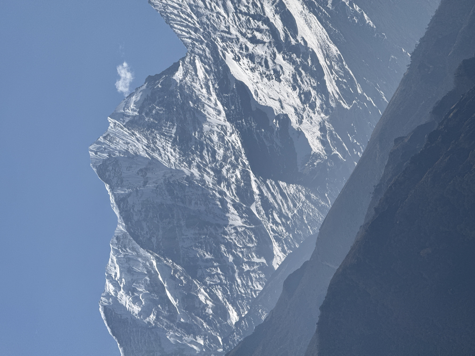 The image captures a breathtaking view of towering, snow-covered mountains under a clear blue sky, characteristic of the Himalayan range near Namche, Nepal. The composition is dynamic, featuring a massive, jagged peak dominating the left side, its steep faces and ridges draped in white snow. The intricate texture of the rock and ice is highlighted by direct sunlight, casting subtle shadows that emphasize the mountain's colossal scale and rugged grandeur.
In the upper left quadrant of the sky, a small, wispy white cloud floats near the mountain's crest, providing a sense of immense height and open space. To the right, successive layers of darker, less-defined mountain slopes recede into the distance, exhibiting strong atmospheric perspective. These slopes appear to be covered in sparse vegetation or rock, rendered in various shades of deep blue and grey due to haze and distance, creating a sense of depth and vastness. The closest dark slope on the far right sharply contrasts with the bright, snow-covered peak, forming a distinct diagonal line across the frame.
The overall impression is one of majestic tranquility and solitude. The clear sky and crisp detail on the main peak suggest good weather conditions, likely during daytime. No specific activity or events are depicted, and there is no visible text in the image.