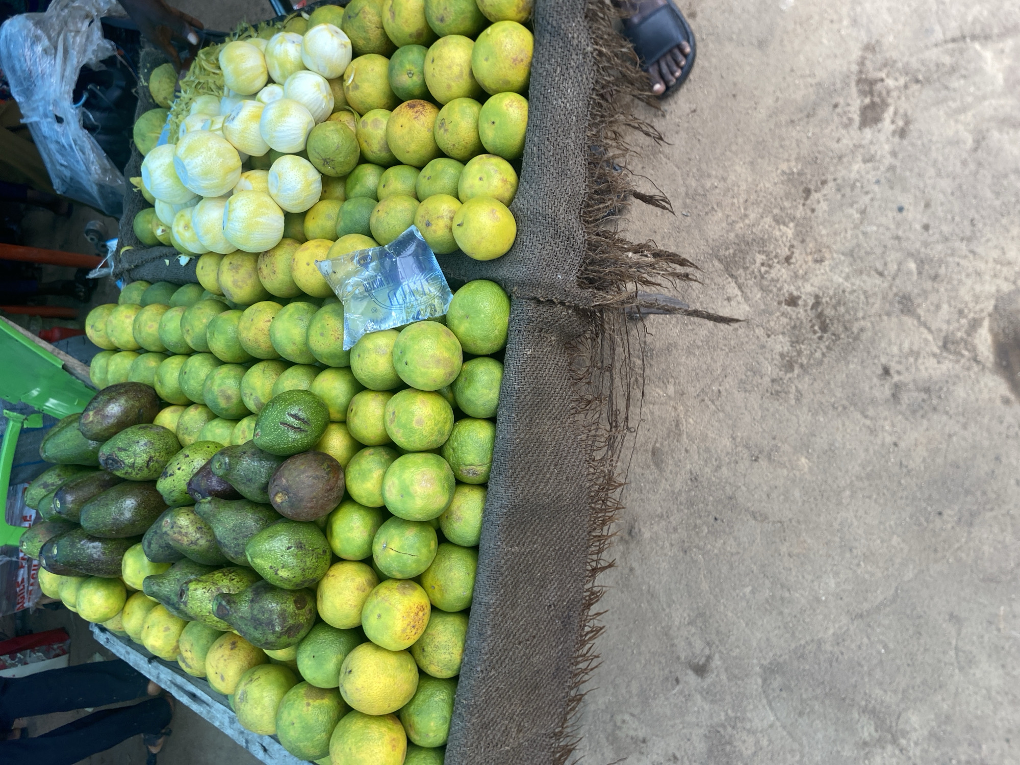 In Jalingo, Nigeria, a market stall displays a generous assortment of fruits, predominantly oranges and avocados. The oranges, varying in shades of yellow and green, are piled high on a coarse brown cloth that drapes over the edge of a wooden table. Avocados, with their distinctive dark green and brownish skin, are intermingled with the oranges, particularly on the left side of the stall. Some of the oranges are quite large, while others are smaller and more uniformly round. A plastic bag containing what appears to be water is visible amongst the oranges. In the background, out of focus, are hints of other market activity and people. The ground beneath the stall is a rough, greyish concrete surface. A person's foot, wearing a dark sandal, is visible at the bottom right of the frame, suggesting an active marketplace. The lighting suggests it is daytime, possibly late morning or early afternoon.