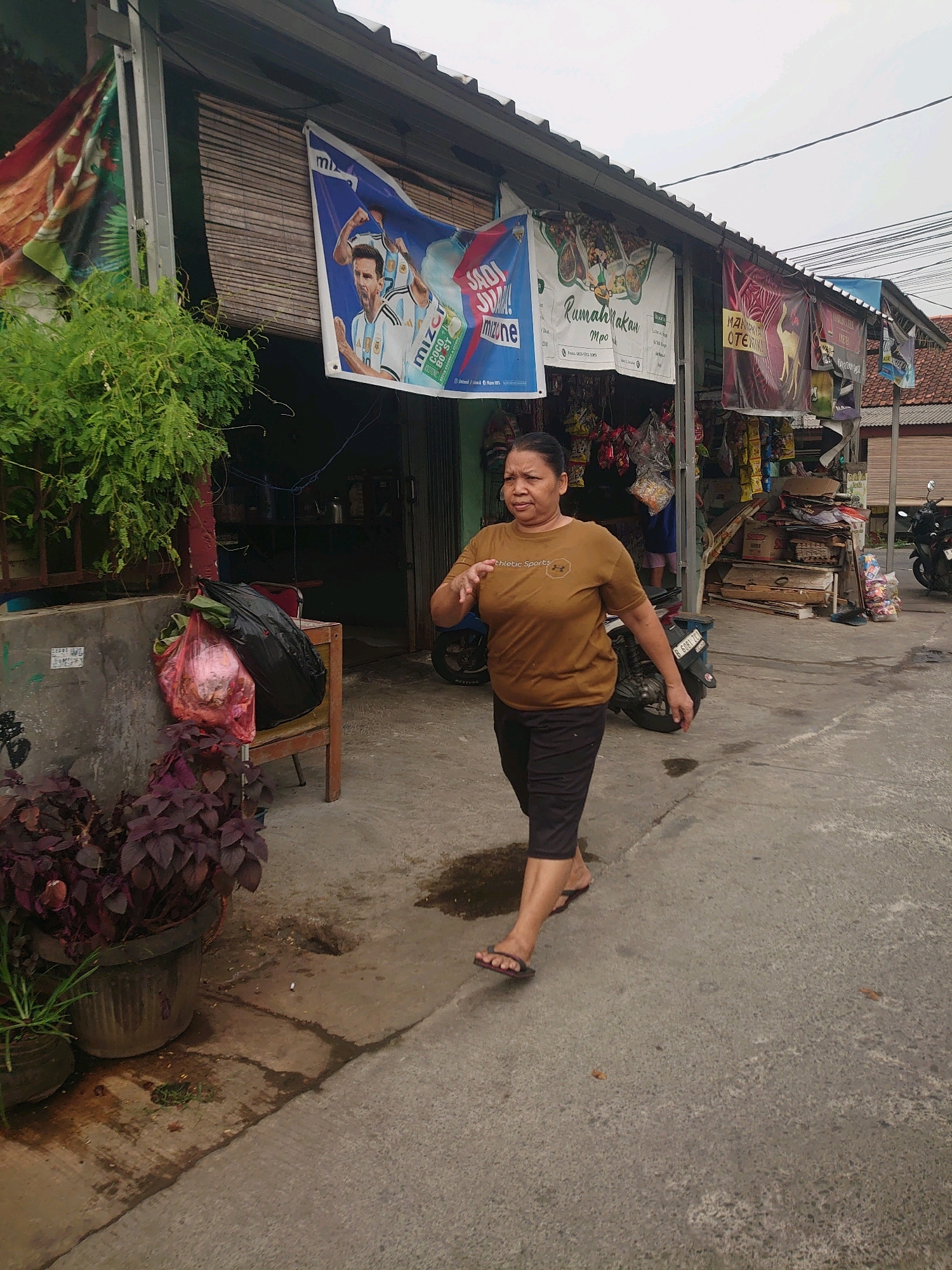 A woman walks on a paved street in front of a storefront in Depok, Indonesia. She is wearing a brown t-shirt with the words "athletic Sports" and black shorts. The storefront has several banners hanging above it, including one with an image of Lionel Messi. There are also motorcycles parked on the street and various items for sale inside the store. The ground is wet in places, indicating recent rain or spillage. There are potted plants on the left side of the image.