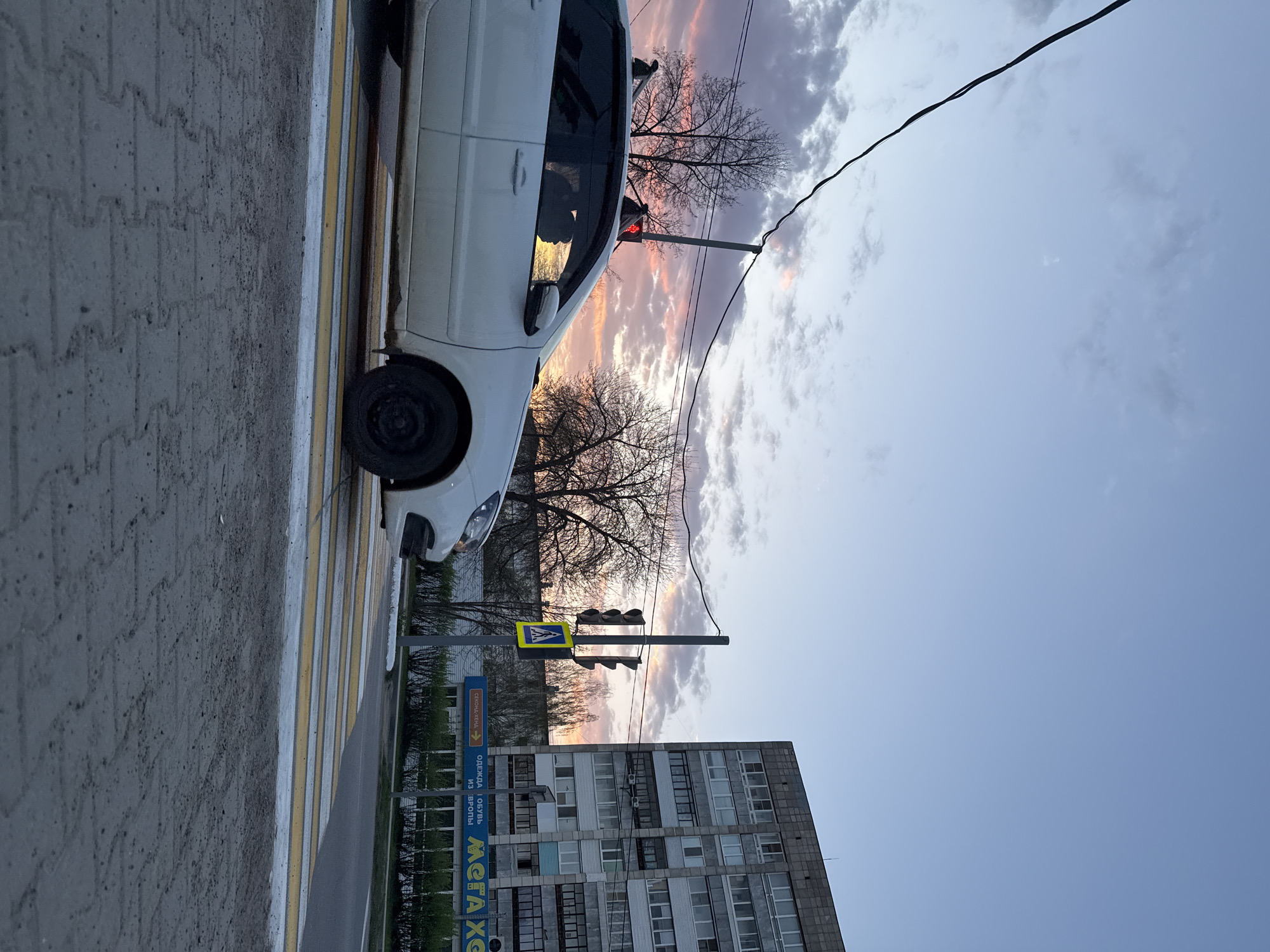 The image depicts a street scene with a white car parked next to a pedestrian crosswalk. The crosswalk has white and yellow stripes. A traffic light pole stands behind the car. A multi-story building is located to the right of the traffic light. The sky is a mix of blue and clouds with hues of pink and orange from a sunset. Bare tree branches are visible against the sky. Overhead wires stretch across the scene. An advertisement sign with Cyrillic text is visible. The location is Unknown, Unknown.