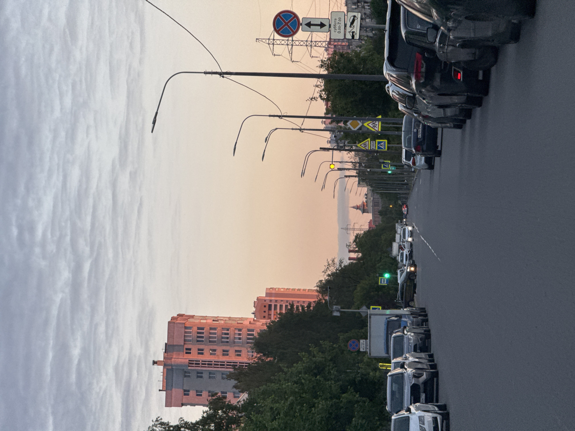 The image depicts an urban street scene during what appears to be late afternoon or early evening, characterized by a soft, warm orange and pink glow on the horizon transitioning to a lighter blue-grey sky with distinct cloud formations.
The main subject is a multi-lane road extending into the distance, lined with numerous cars. A row of vehicles is parked along the right side of the road, facing away from the viewer. Further down the street, additional cars are visible, suggesting traffic or parked vehicles.
Several streetlights with curved poles illuminate the road. A multitude of road signs are present, including a circular blue regulatory sign with a red cross (indicating "No Stopping" or "No Parking"), beneath which Cyrillic text reads "Суббота 0:00-7:00" (Saturday 0:00-7:00). Other visible signs include directional arrows, yellow diamond-shaped warning signs (one indicating "100m"), and a green traffic light.
To the left of the road, behind a line of green trees, stands a prominent multi-story building with a reddish-brown facade and numerous windows. Other buildings and dense foliage are scattered along the street in the background. A large metal electricity pylon with power lines is also visible in the mid-ground.
The overall scene portrays a typical urban environment, likely in Vsevolozhsk, Russia, with no specific activity beyond the routine flow of traffic and parked cars under a serene, partly cloudy sky.