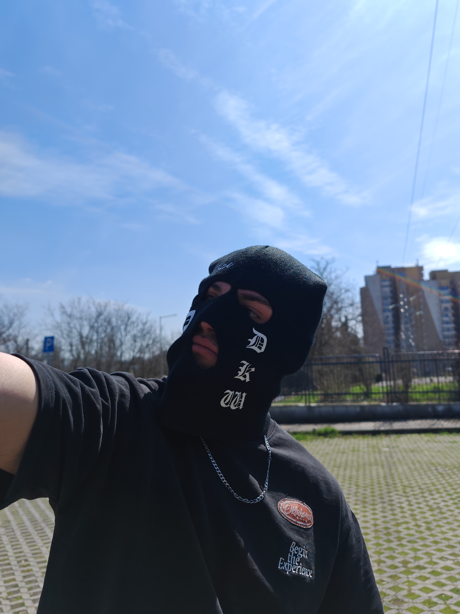 The image depicts an individual in an outdoor, urban setting within Shumen, Bulgaria, under a clear blue sky with scattered cirrus clouds. The subject, captured from the chest up, wears a black balaclava featuring white gothic-style lettering, possibly "D K W", across the forehead and left cheek. The individual's eyes and the area around the mouth are visible through the balaclava's openings. A black short-sleeved t-shirt is worn, accessorized with a silver-colored chain around the neck. On the right chest of the t-shirt, an oval patch with "Oklahoma" text is present, above "Begin the Experience" embroidered text. The individual's right arm is extended, holding the camera for a self-portrait.
The background shows a paved ground surface composed of interlocking concrete pavers with interspersed grass. This transitions to a dark, horizontal metal fence or railing. Beyond the fence are several bare deciduous trees, indicating a season of dormancy. In the far background, a multi-story residential building, consistent with Eastern European apartment block architecture, stands under the bright sun. A blue square parking sign with a white 'P' is visible in the mid-ground on the left. Faint power lines traverse the upper right portion of the sky, and a subtle lens flare is visible near the residential building.
