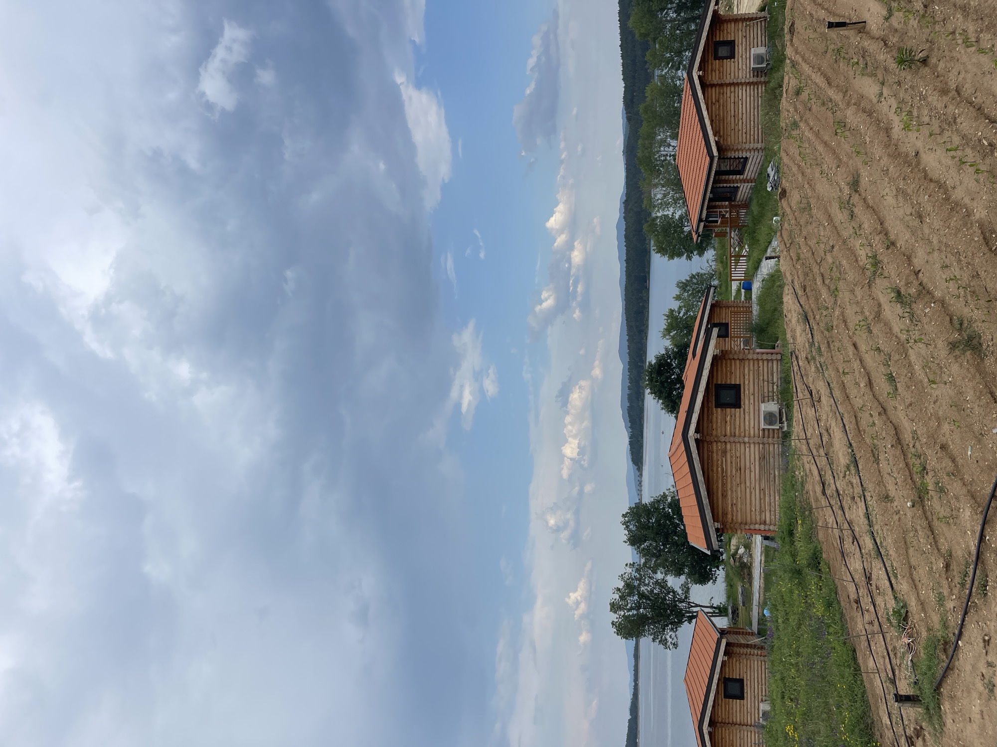The image depicts a rural landscape near the Koprinka Reservoir in Koprinka, Bulgaria, under an overcast sky with patches of blue. In the immediate foreground, a sloping field of reddish-brown tilled earth is organized into parallel rows. Small, green seedlings are visible in these rows, irrigated by black plastic hoses running along the ground. To the left of the tilled field, an area of wilder green grass and scattered yellow and purple wildflowers is present.
In the mid-ground, three wooden log cabins with dark-stained window frames and bright orange/terracotta-tiled gable roofs are visible. The central cabin is fully visible, featuring a window on its side and an outdoor air conditioning unit mounted below it. A wooden railing suggests a deck or porch facing the water. To its immediate left, another cabin is partially visible. A third cabin is seen on the far right, also with an outdoor air conditioning unit. These cabins are situated on a grassy area, transitioning down towards the water.
The background is dominated by a large, calm body of water, likely the Koprinka Reservoir, stretching horizontally across the scene. The distant shore is lined with green trees. Beyond the water, rolling hills or mountains, covered in vegetation, appear bluish in the distance. A faint structure, possibly a bridge or part of a dam, can be discerned spanning the water on the right side of the background. The sky above is largely covered by grey, heavy clouds, particularly in the upper frame, but clears towards the horizon to reveal patches of light blue sky with scattered white cumulus clouds. The lighting is diffused, indicating an overcast day.