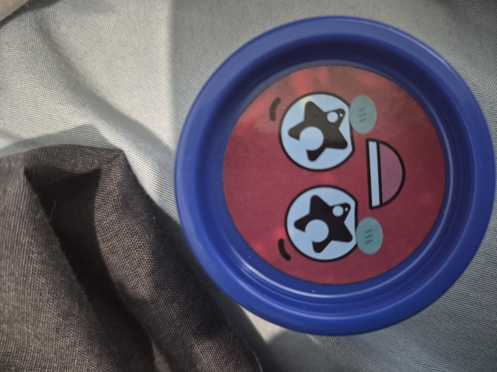 The image captures a close-up, top-down view of a circular object featuring a playful, cartoon-like face, resting on textured fabric. The main subject is a blue-rimmed circular item, likely plastic, with a red center where the face is depicted. This face has two prominent light blue, circular "eyes," each containing a black, star-like pupil with a white dot for a highlight. A wide, red mouth with a white interior is visible on the right side of the face, suggesting an open or surprised expression. Small, black curved lines near the eyes contribute to the animated character. A small, light green oval-shaped detail, possibly a sticker or printed element, is positioned between the right eye and the mouth, clearly displaying the number "111."
The object is situated on two distinct fabric surfaces. The background on the right and top consists of a light-colored, possibly light blue or grey-blue, woven fabric with a visible, fine texture. On the left and bottom, a darker, likely dark grey or black, fabric is seen, appearing folded and creating soft shadows. The scene is evenly lit, indicating an indoor setting or diffused outdoor light. There are no people present, and no specific activity is taking place. The object itself appears to be a lid, a coaster, or a small toy, possibly associated with children's items. The scene is set in Yerevan, Armenia.