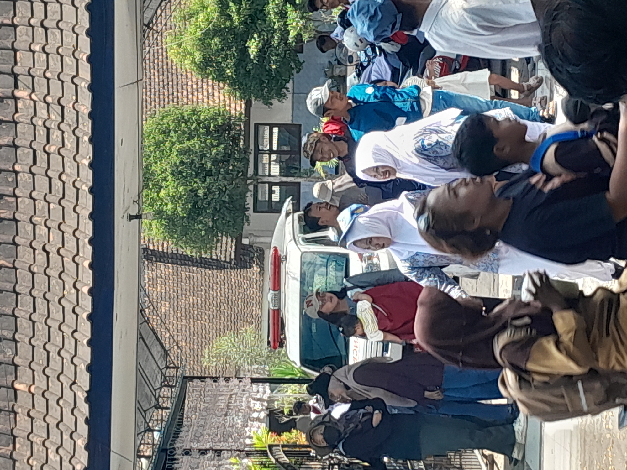 In Genteng, Indonesia, a crowd of people, some wearing hijabs, gather outdoors under a tiled roof. A white vehicle with red flashing lights, possibly an ambulance or police car, is visible behind them. Some individuals are carrying bags, and a child is being held by an adult. The environment appears to be a sunny day, with trees in the background.