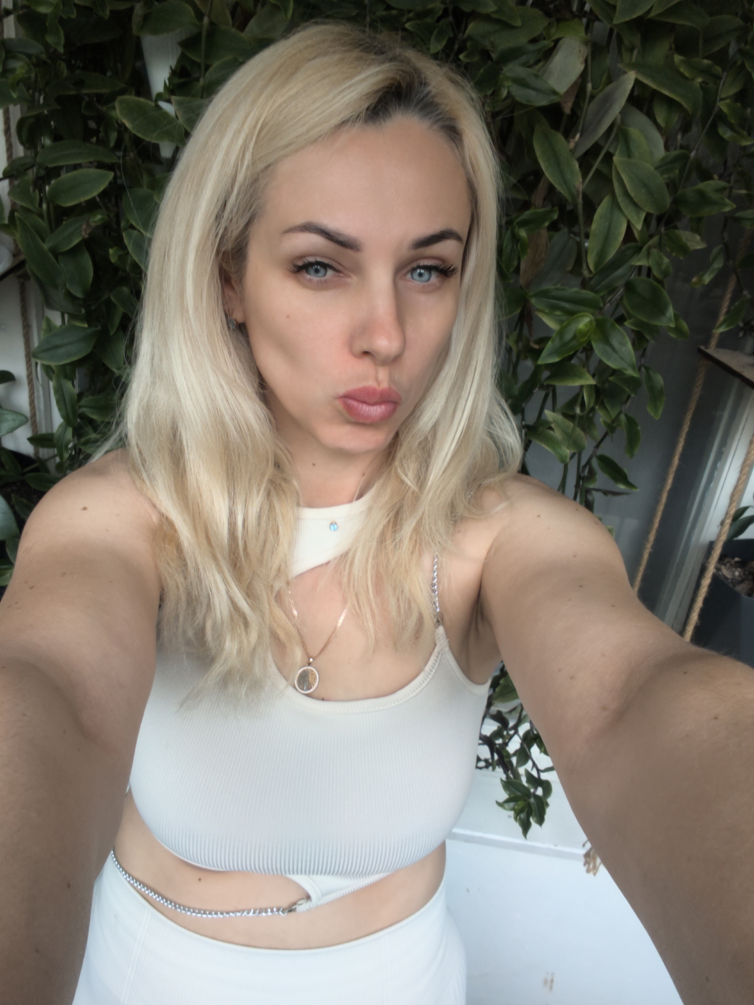 A fair-skinned, blonde-haired woman is taking a selfie. She has blue eyes and pursed lips. She is wearing a light-colored ribbed tank top with a cutout detail at the collarbone and a silver chain belt around her waist. A necklace with a round pendant is visible. The background features green leafy plants. A window with a rope hanging down is visible to the right. It is not possible to identify the location as Sevastopol, Ukraine, or to provide any additional information about actions or interactions based on this single image.