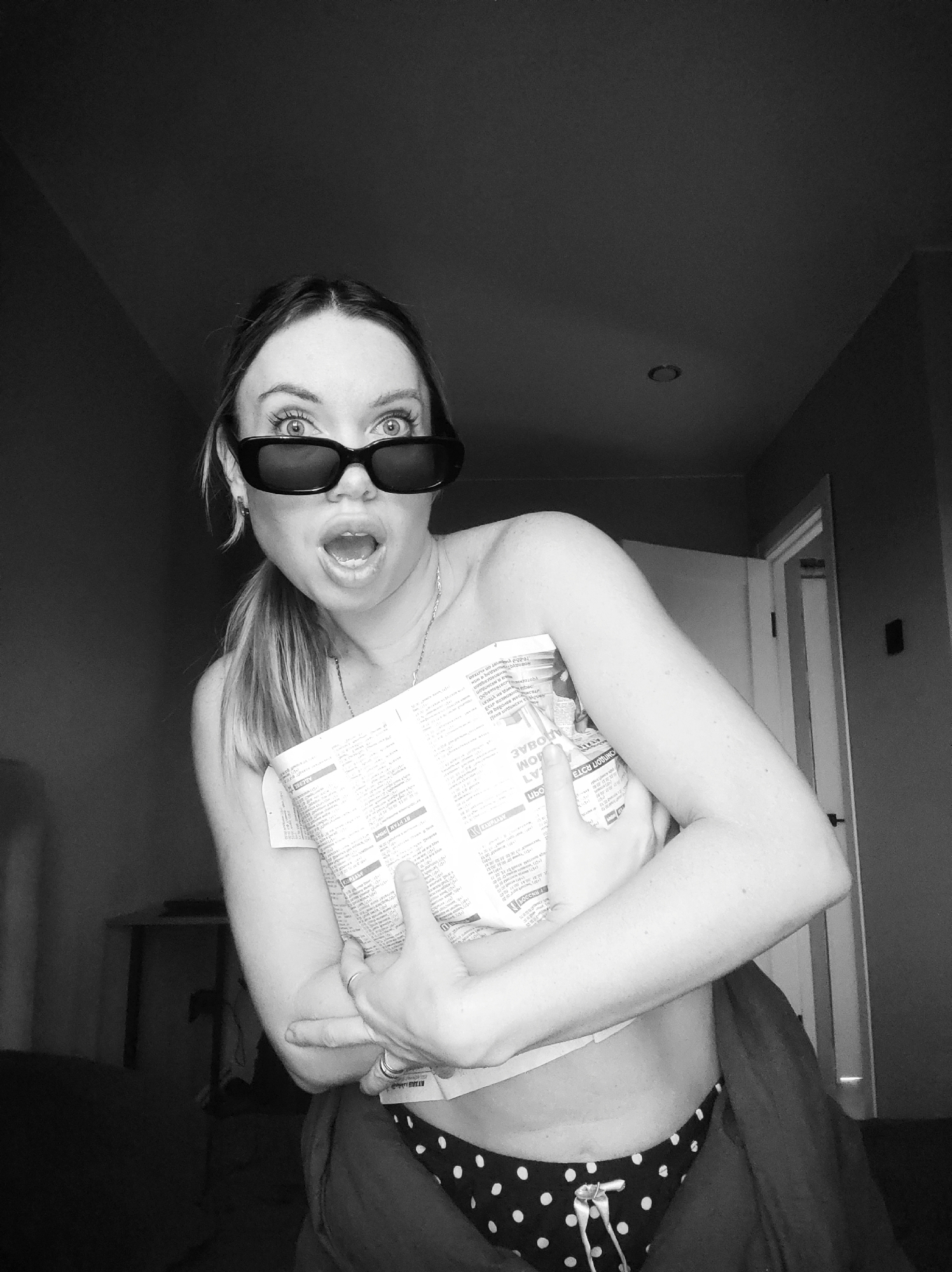 A black and white photograph captures a young woman indoors in what appears to be a bedroom or living space in Votkinsk, Russia. She is the central subject, looking directly at the camera with a dramatic expression of surprise or shock, her eyes wide above dark, rectangular sunglasses and her mouth open. Her hair is styled in a ponytail. She is wearing a light-colored top, possibly strapless, and black polka-dot bottoms with a white drawstring tie. Two rings are visible on her left hand.
She holds a crumpled newspaper or brochure to her chest, which has mirrored text that is mostly illegible but shows fragments like "MOM," "LY," and "BETAE." The background is dimly lit, featuring dark walls, a partially open white door on the right, and a circular recessed light fixture on the ceiling. A table or desk is faintly visible on the left side of the frame. The overall atmosphere is candid and suggests a moment of humorous astonishment.