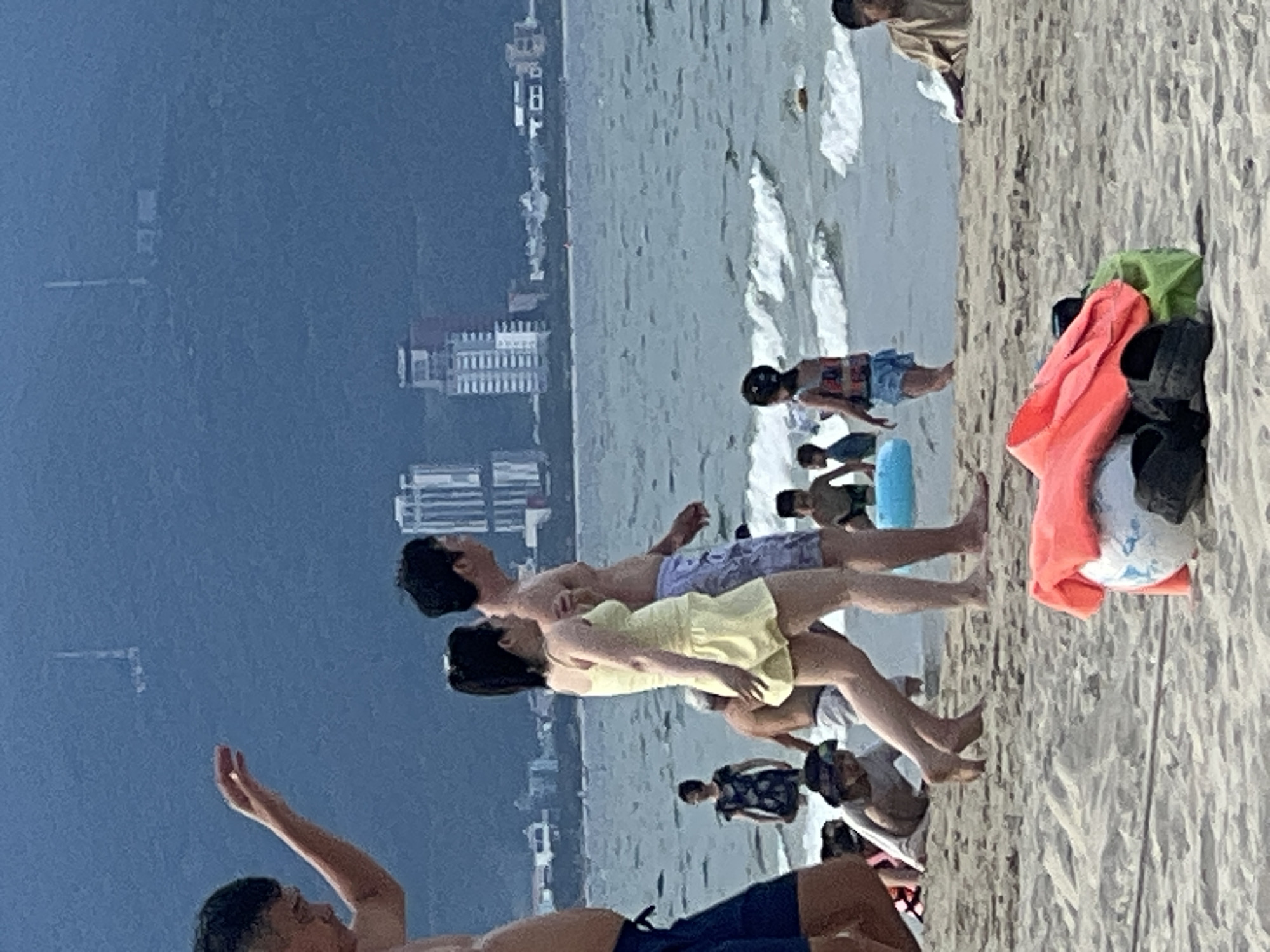 The image depicts a crowded beach scene in Đà Nẵng, Vietnam. The background features a hazy view of the city skyline with several high-rise buildings, including what appears to be a hotel or residential complex under construction. The sea is visible in the middle ground with moderate waves. On the beach in the foreground, several people are present. A couple in the center is walking away from the camera, with the woman wearing a yellow dress and the man in shorts. To the left, a man's arm is raised, and he is partially visible. Other individuals, including children, are scattered across the sand and in the shallow water. In the immediate foreground, beach gear, including a life jacket, a ball, and sandals, are piled on the sand. The lighting suggests it might be late afternoon or early evening, with overcast conditions contributing to the muted colors.