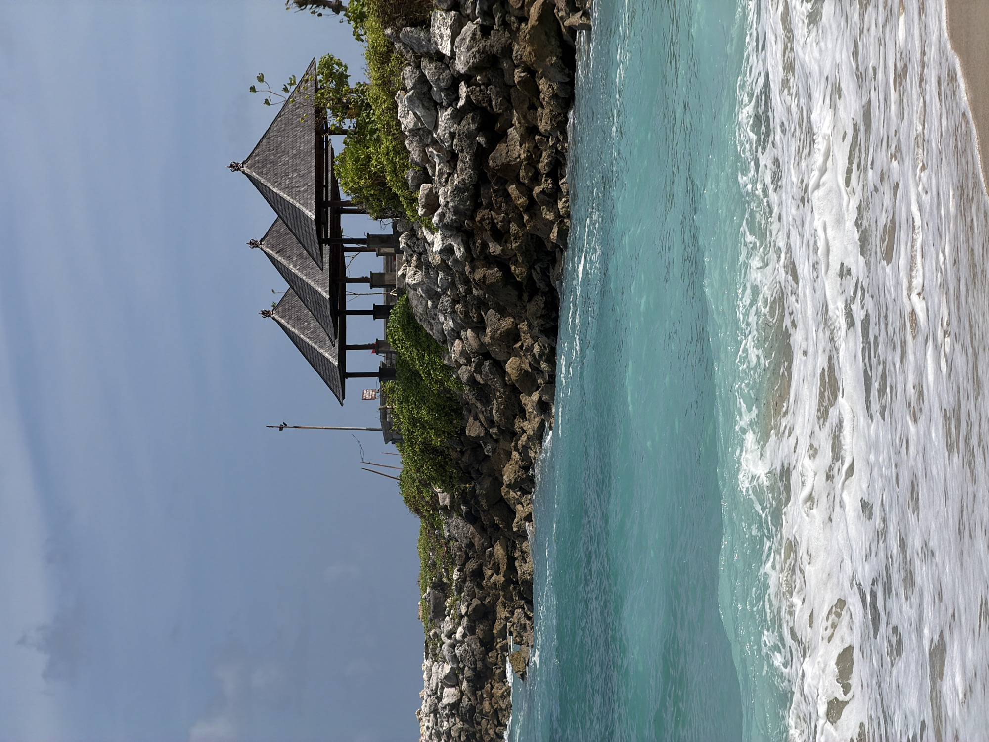 The image presents a view of the shoreline in Kuta Selatan, Indonesia, with a distinctive structure perched on a rocky cliff. The turquoise water of the ocean is visible, edged with foamy waves washing onto the sand. The structure on the cliff appears to be a gazebo or small pavilion with tiered roofs, possibly a traditional Balinese design. The cliff itself is composed of rocks and is partially covered with green vegetation. In the background, a cloudy sky can be seen.