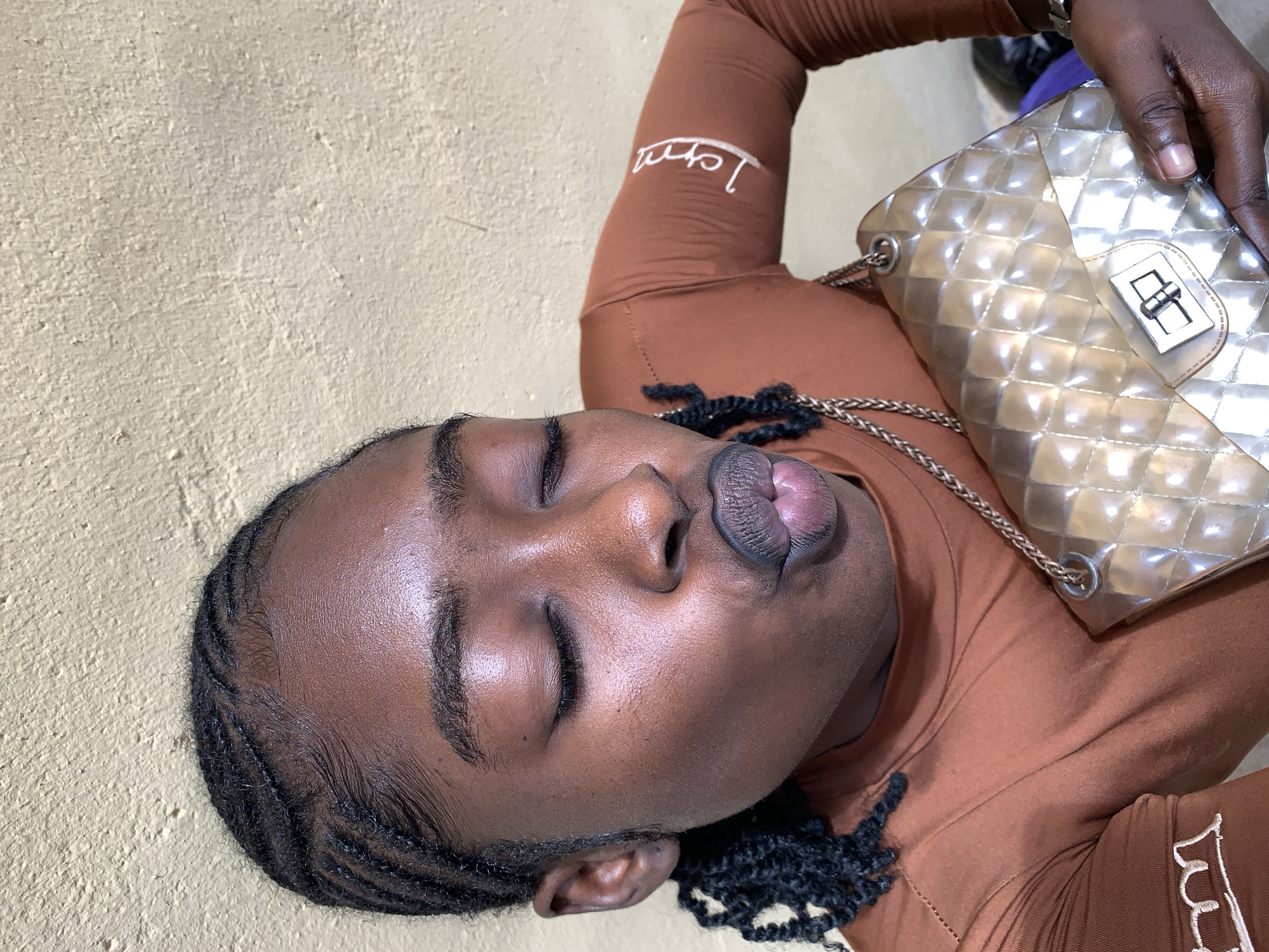A young Black woman is depicted in Rafin Par, Nigeria, posing with her eyes closed and lips pursed into a "kissing" or "pout" expression. Her hair is styled in cornrows with neat baby hairs along the hairline, and some braided sections hang down. She wears a form-fitting, reddish-brown long-sleeved top featuring white stylized text, possibly "Love," embroidered on the left forearm. The woman holds a quilted, light gold or iridescent PVC/patent leather handbag with a metallic turn-lock closure and a chain strap. A silver-toned watch with a link bracelet is visible on her left wrist. The background consists of a textured, light beige or yellowish-brown wall, indicating an outdoor or brightly lit indoor setting.