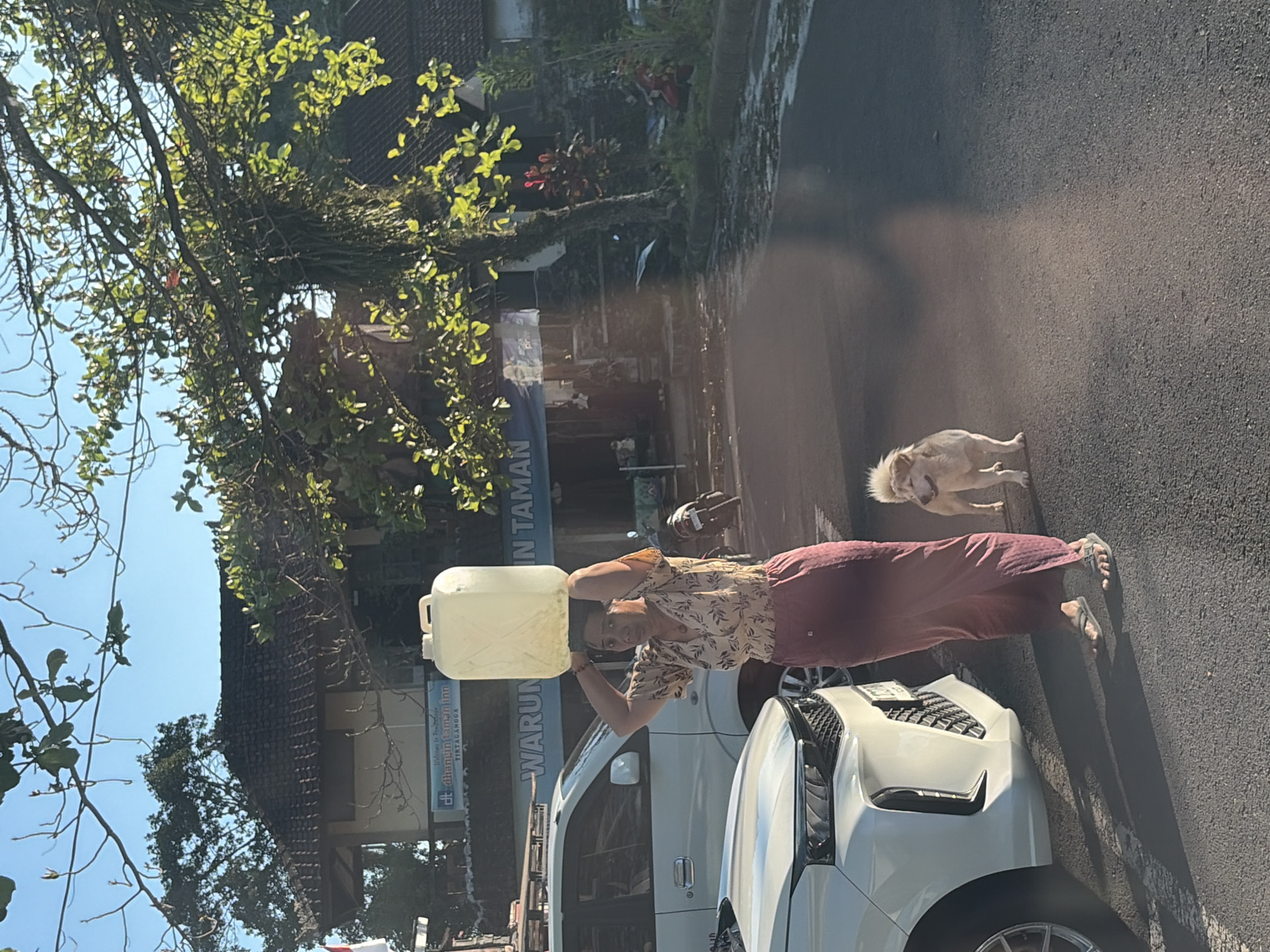 In Abang, Indonesia, a woman carries a large, translucent plastic container of water balanced on her head, walking on a paved street. She is wearing a floral print blouse and a long, maroon sarong. A small, light-colored dog stands next to her. In the background, a white SUV and a motorcycle are parked. A building with a tiled roof and a sign that reads "WARUNG IN TAMAN" is visible. Lush green trees and vegetation surround the area, and the sky is clear blue. The sunlight casts long shadows on the ground, indicating it is likely morning or late afternoon.