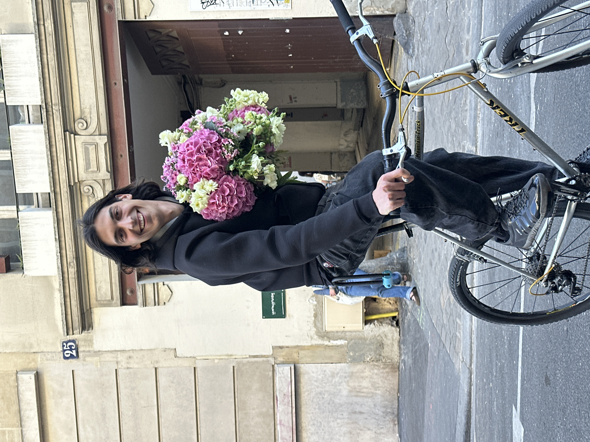 A man on a bicycle holds a bouquet of flowers outside a building with the number 25 on the wall. The bicycle is silver with black tires, and the man is wearing a dark jacket and pants. The flowers are a mix of pink and white. The scene appears to be in Paris, France, as indicated by the architectural style. Other people are visible in the background walking down the street.