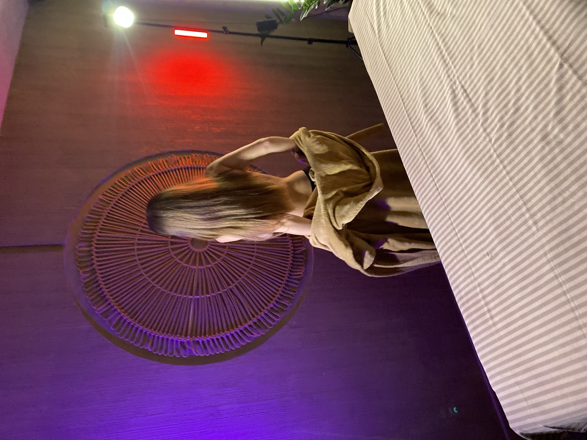The image depicts a woman seated on a wicker chair, partially covered with a brown robe or blanket. The chair is placed against a wall, illuminated with purple lighting from below. Above, the wall has a reddish glow. A striped bed or fabric is visible on the right side of the frame. Above the red glow on the wall is a white light. The location in Izhevsk, Russia, is not deducible from the provided image.