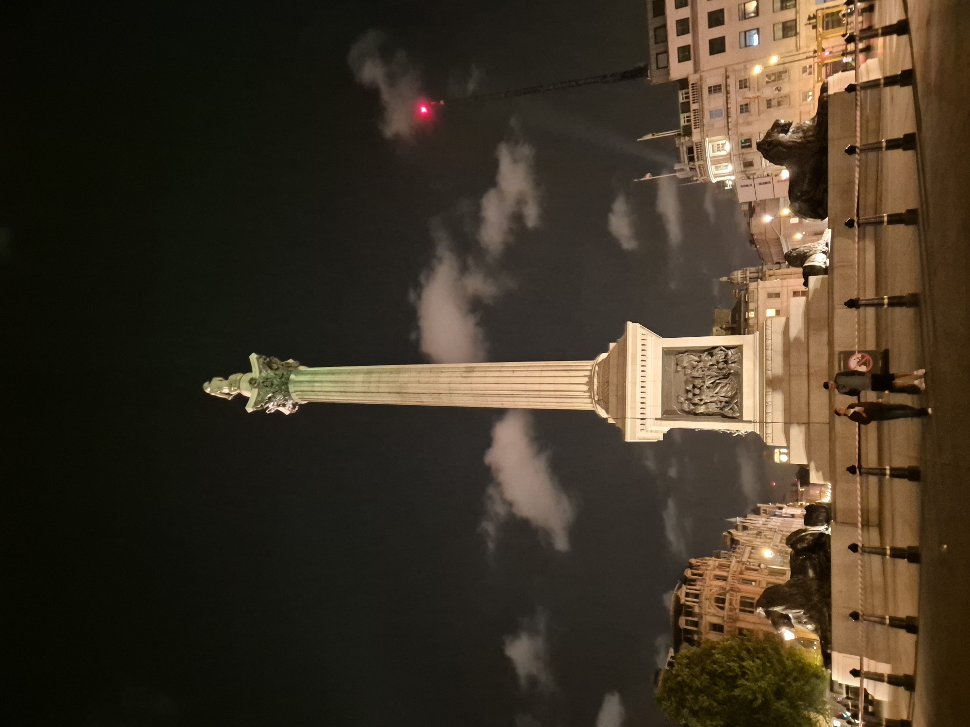 This is a nighttime, low-angle, wide shot of Nelson's Column in Trafalgar Square, London, United Kingdom. The iconic monument, topped by a statue of Admiral Lord Nelson, is illuminated against a dark sky with scattered clouds. At the base of the column are reliefs depicting naval battles. To the left and right of the column are large bronze lion sculptures. Two people, a man and a woman, are standing near the base of the monument, facing away from the camera. In the background, traditional London architecture with lit windows can be seen, along with a tall radio mast with a red blinking light. The scene is cordoned off with white tape and black stanchions.