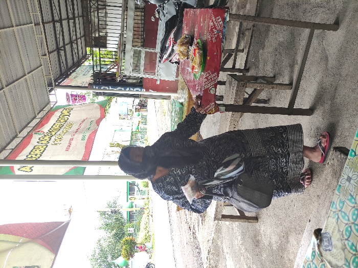 An outdoor scene in Kepanjen, Indonesia, captures a woman interacting with what appears to be a street stall or small shop. The image is taken during the daytime under bright conditions.
A woman, wearing a dark, patterned long dress and a black head covering (hijab), stands on a concrete surface. She has a grey shoulder bag slung across her body and is wearing pink flip-flops. Her right arm is extended, reaching towards items displayed on a table to her right, suggesting she is selecting or arranging snacks.
The table in front of her is rustic, possibly made of wood or metal, with a red surface. It holds several packaged snacks, such as chips or crackers, some arranged on a small green tray. A simple wooden stool or chair is placed under the table.
The entire area is covered by an open-sided metal roof structure, providing shade. In the background, a large banner stretches horizontally, displaying text and patterns. Beyond the banner, an outdoor vista reveals distant buildings, lush green trees, and a bright sky. A portion of a motorcycle or scooter is partially visible on the far right.
Visible text on the banner includes Indonesian phrases such as "CABUTAN BERHADIAH KOTAN," "KAPANAN... SELAWA... JAYA..." and "SELATAN KOTA MALANG" (South Malang City), as well as "JUMLAH PELATIH" (Number of Trainers). The word "KAPANAN" likely refers to Kepanjen, aligning with the provided location context.