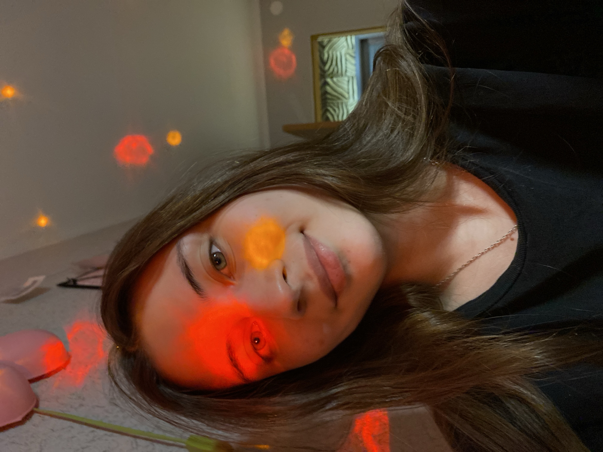 A young woman with long brown hair is lying on a light-colored surface. She is wearing a black shirt and a silver necklace. Her face is illuminated with colored lights. Red lights are projected onto her left eye and cheek, and an orange light is projected onto her nose. There are more red and orange lights projected onto the background wall and ceiling. In the background, there is a framed picture on the wall. In the bottom left corner, there are pink balloons on a table.
There is no clear indication of this being located in Votkinsk, Russia.