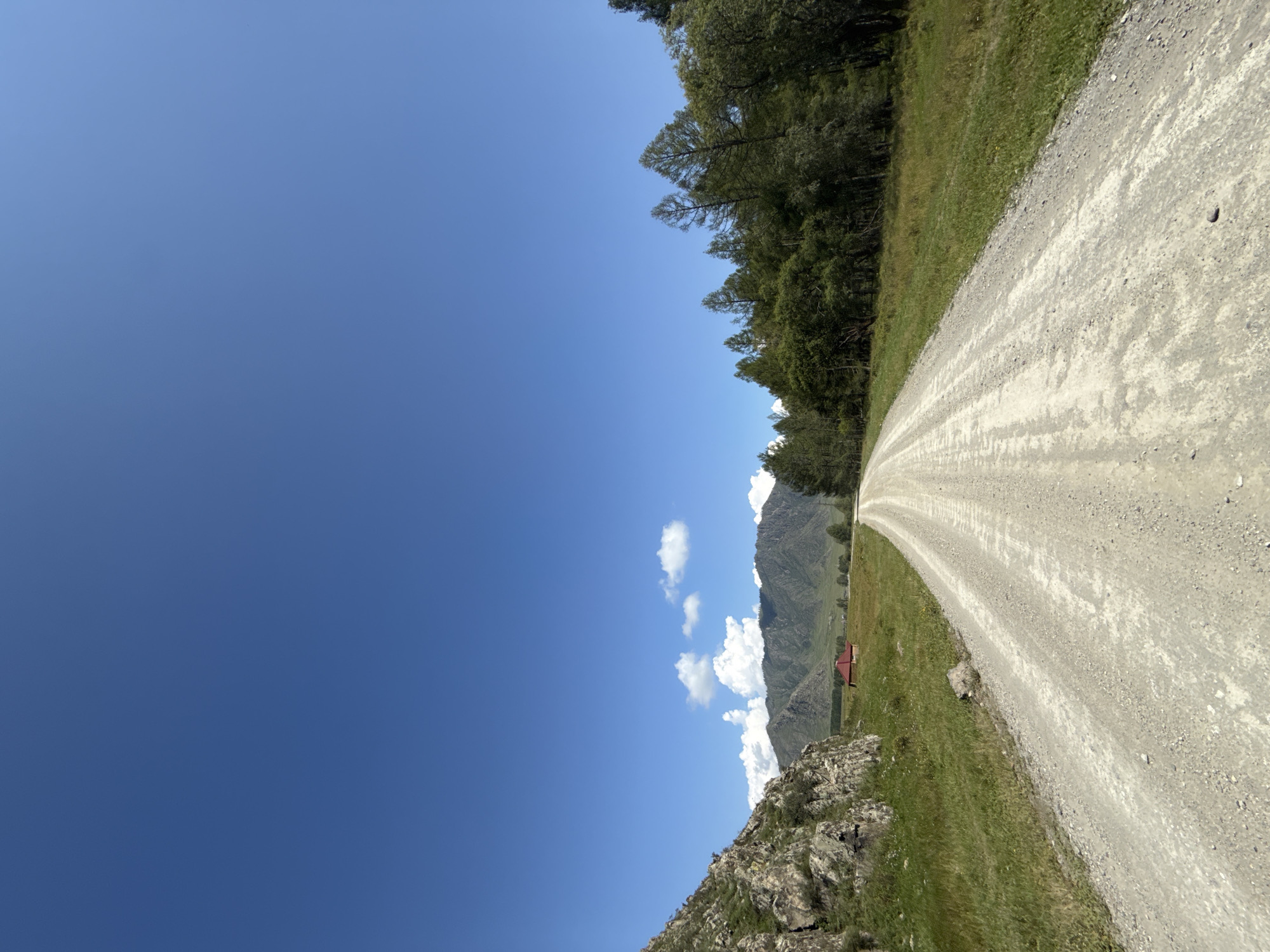 A bright, sunny day in Bichiktu-Boom, Russia. The image shows a dirt road stretching into the distance, flanked by green grassy fields. On the left side of the road, a rocky, moss-covered hillside rises. In the middle distance, a small, red-roofed building sits in a field. Behind the field, a range of majestic mountains are visible, their peaks partially obscured by fluffy white clouds against a brilliant blue sky. On the right side of the road, a dense forest of evergreen trees creates a dark, verdant border. The road itself is wide and appears to be made of light-colored gravel and dirt, with tire tracks visible. The scene conveys a sense of open space, natural beauty, and rural tranquility.