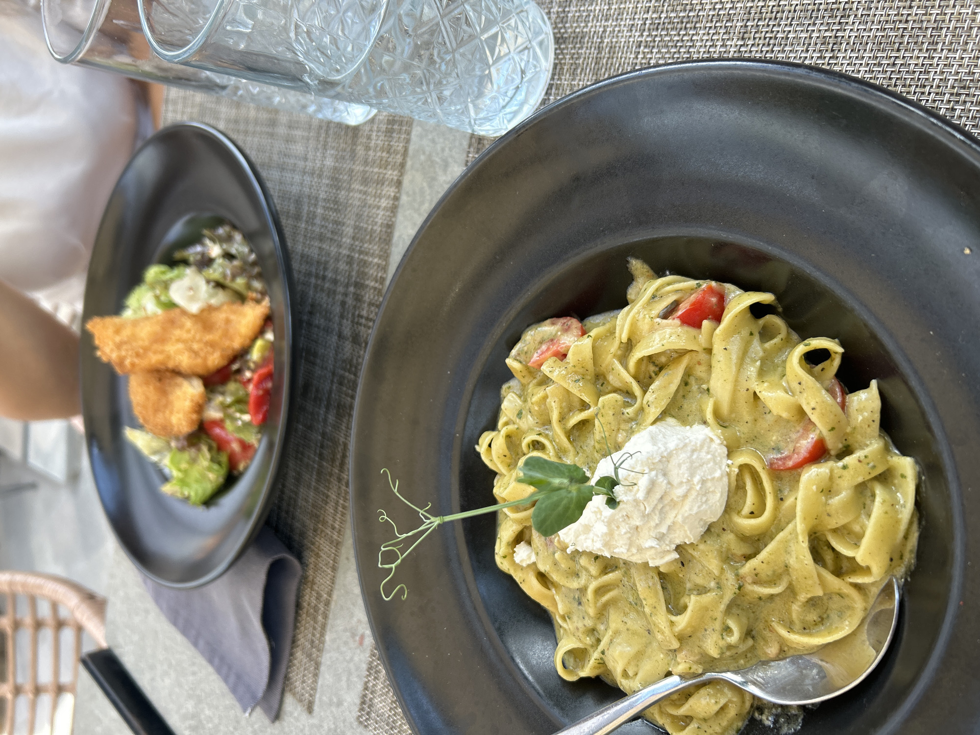 The image shows a table setting with two black plates containing food. On the left plate is a salad with fried cheese and tomatoes. On the right plate is a pasta dish with a dollop of white cheese on top, garnished with a pea shoot, and a spoon. There are two drinking glasses in the background. The table has a woven pattern. Based on the style of food and the presence of Bulgarian currency, the location is likely Sofia, Bulgaria. There is also a partial view of a person's arm wearing a light-colored shirt on the left side of the image.
