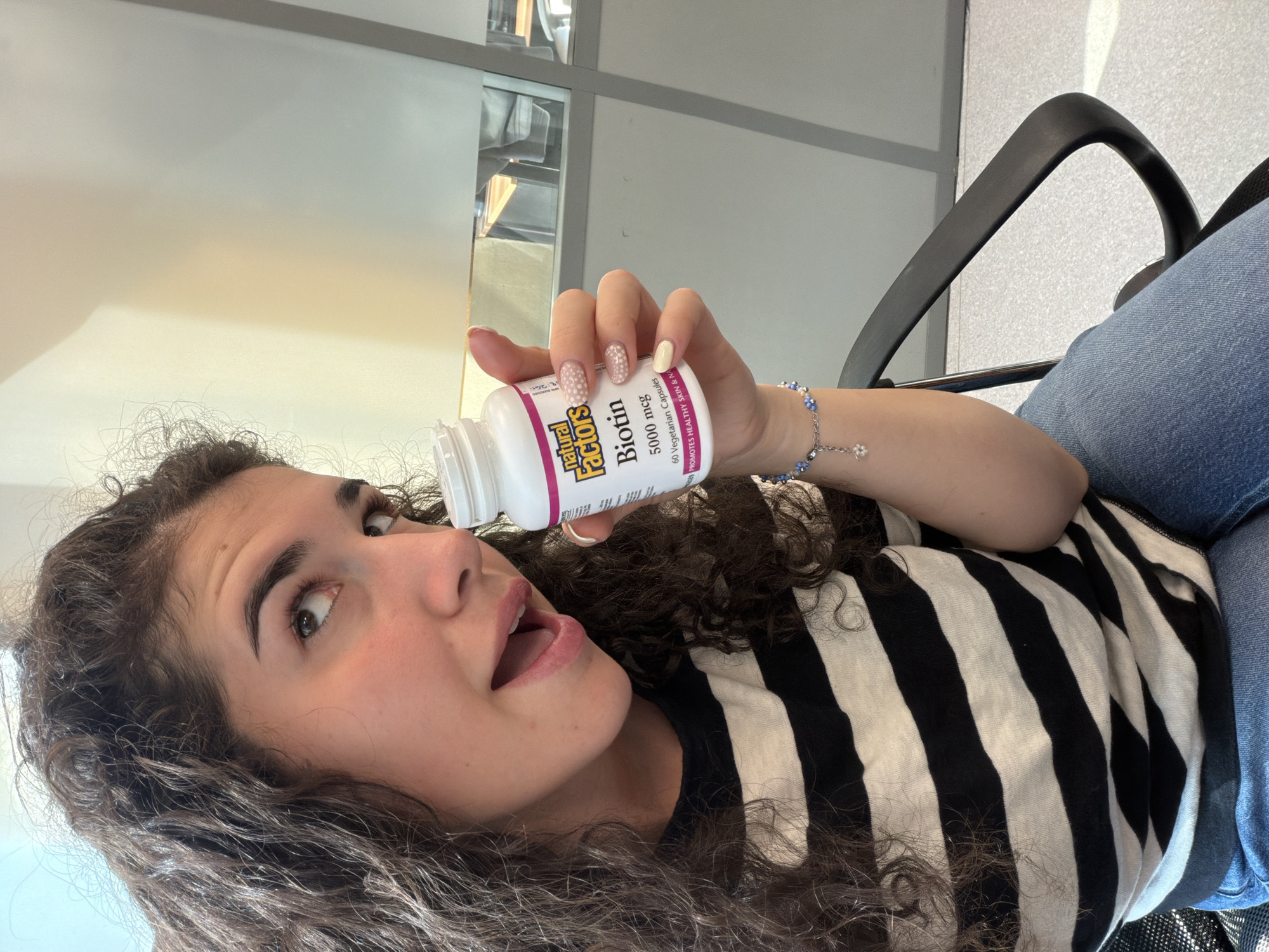 A young woman with curly brown hair, wearing a black and white striped shirt and blue jeans, sits in a black office chair. She holds a white bottle of Natural Factors Biotin supplements in her right hand. Her mouth is open, and she is looking up and to the left. The background appears to be an indoor setting with a light-colored wall and a frosted glass partition. There is no visible indication of the location being in Sofia, Bulgaria.