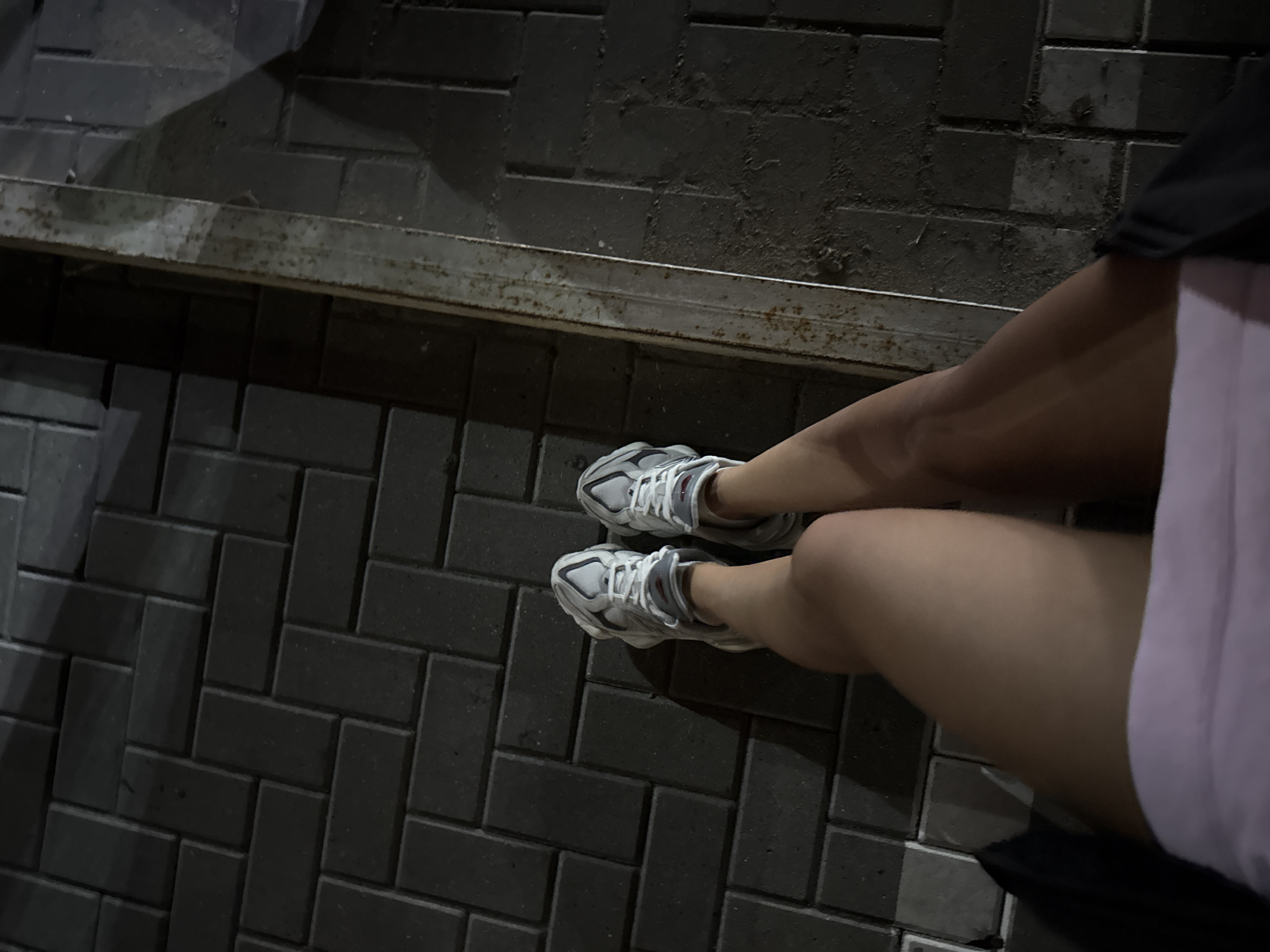 This is a high-angle, full-body shot of a person's legs from the knees down, wearing white athletic shoes and a light pink garment. The person is standing on a dark, patterned brick walkway, possibly in Cimișlia, Moldova. To the right of the person's legs is a metal divider strip, separating the brick walkway from another surface which is out of focus. The lighting suggests it is nighttime or a dimly lit area, casting shadows from the person and their shoes onto the ground.