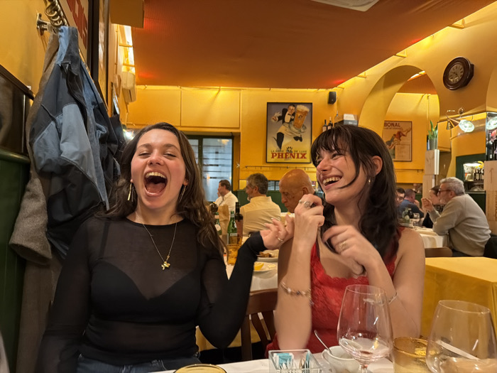 Two young women are sitting at a table in a restaurant, laughing. The woman on the left is wearing a sheer black top and has her mouth wide open. The woman on the right is wearing a red top. There are wine glasses on the table. In the background, there are other patrons and posters on the yellow walls. A coat hangs on the left of the shot.