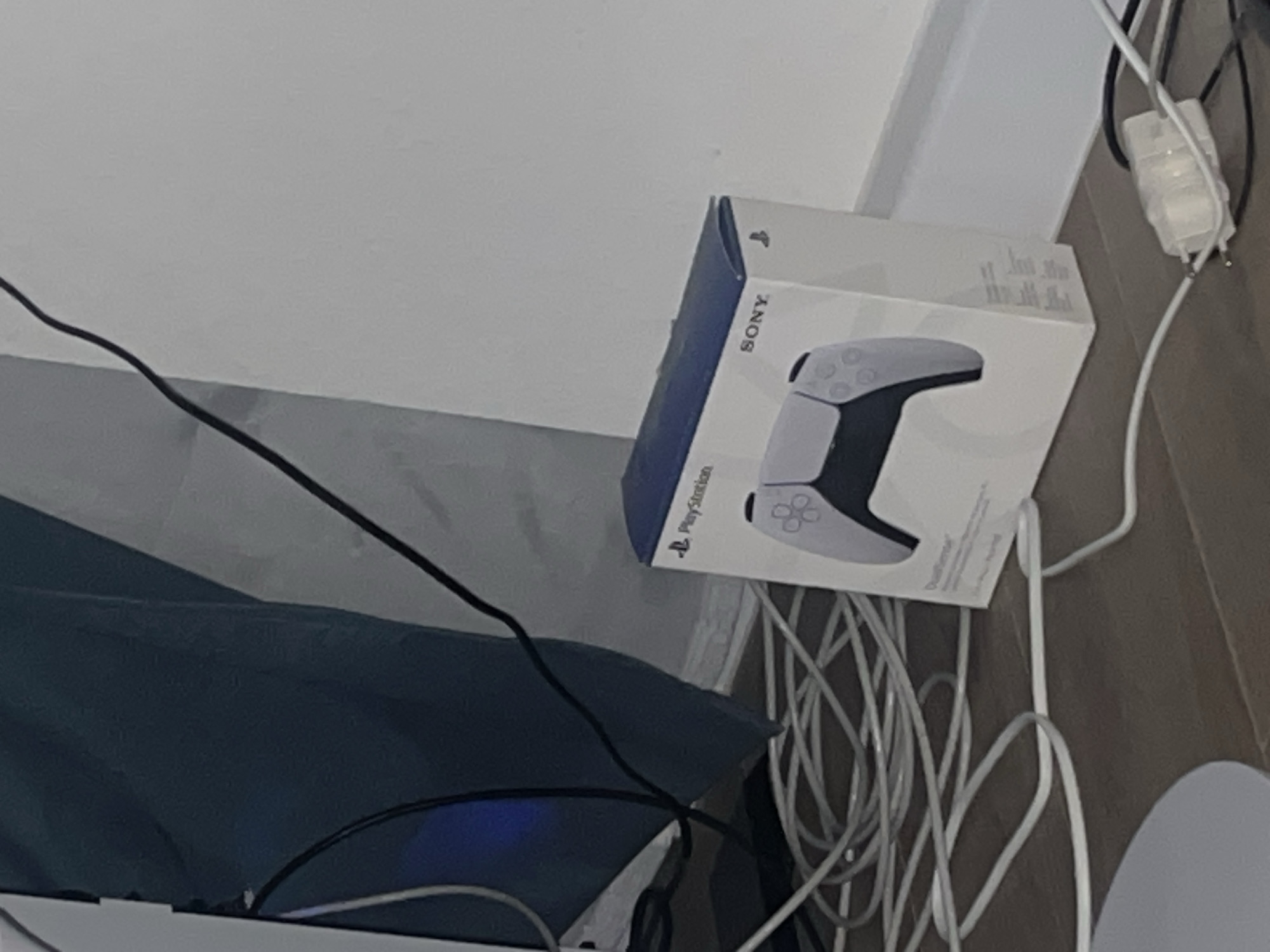 The image shows a box for a PlayStation DualSense controller placed on a floor next to a bundle of white and black cables. The box is white with blue accents and features an image of the controller. A dark blue fabric is visible in the background, along with a white wall. The scene appears to be indoors, possibly in a room, with no specific urban landmarks identifiable. The lighting is somewhat dim, casting shadows on the floor. There are no people or animals present in the image. The cables appear to be connected to some electronic device, possibly a gaming console or computer, which is not fully visible. The overall impression is that of a casual, domestic environment with gaming equipment.