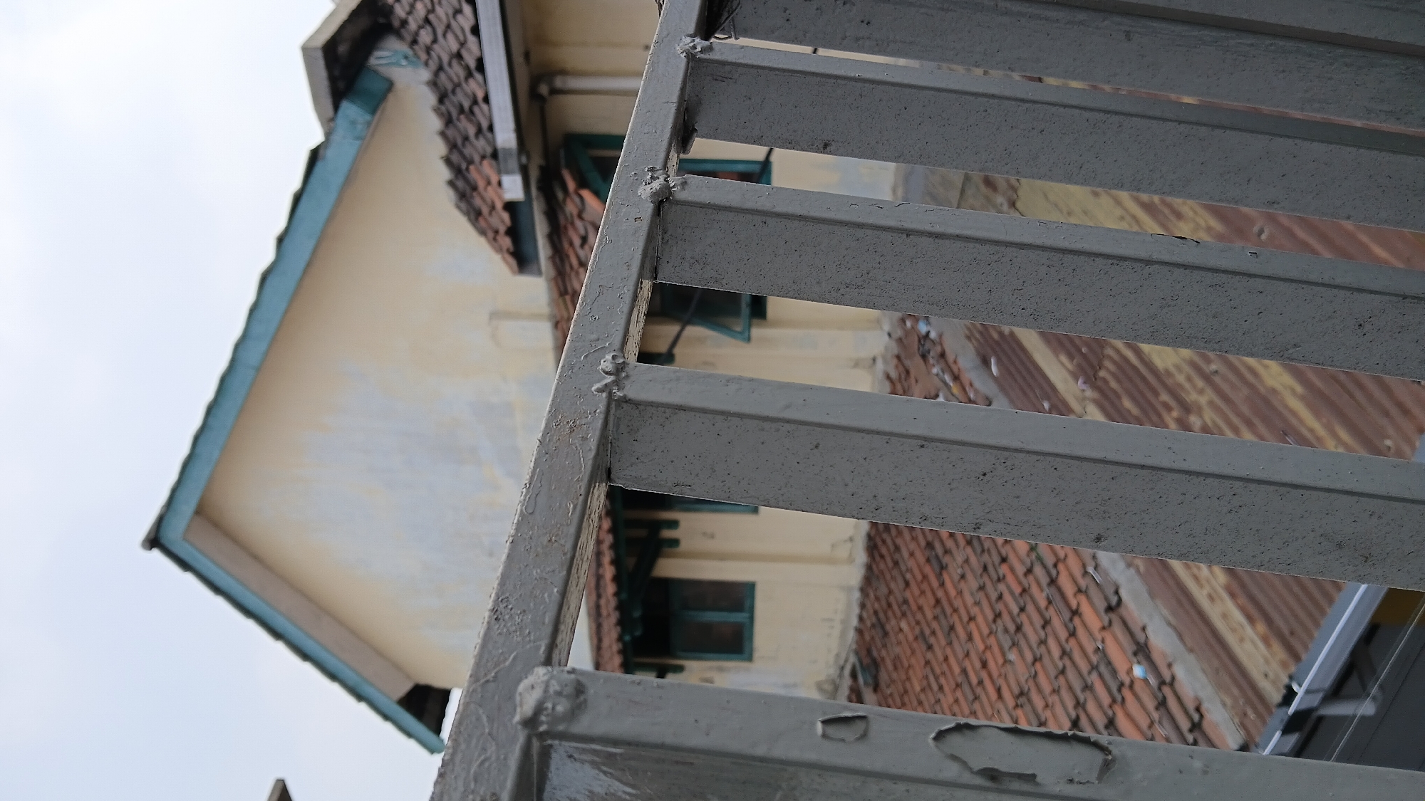 The image depicts a slightly overcast sky above a building's roofline, viewed through a gray metal railing. The railing, made of vertical bars and a horizontal top section, occupies the foreground and is textured with specks and peeling paint. Beyond the railing, the building features a light-colored facade with a turquoise-trimmed roof and several terracotta-tiled sections. Visible windows with turquoise frames are set into the wall. Below the main roof, corrugated metal roofing and more terracotta tiles are apparent. The scene suggests an urban or suburban environment, potentially in Bandung City, Indonesia, given the architectural style and materials. No people or specific actions are observable.
