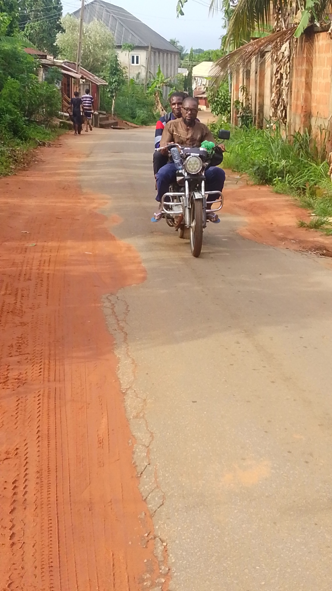 Two men are riding a motorcycle on a paved road in Enugwu-Ukwu, Nigeria. The road is partially covered with red soil. The man in front wears a brown shirt and glasses and is operating the motorcycle. The passenger behind him is wearing a striped shirt. In the background, other people are walking on the side of the road, and there are buildings and vegetation lining the road.