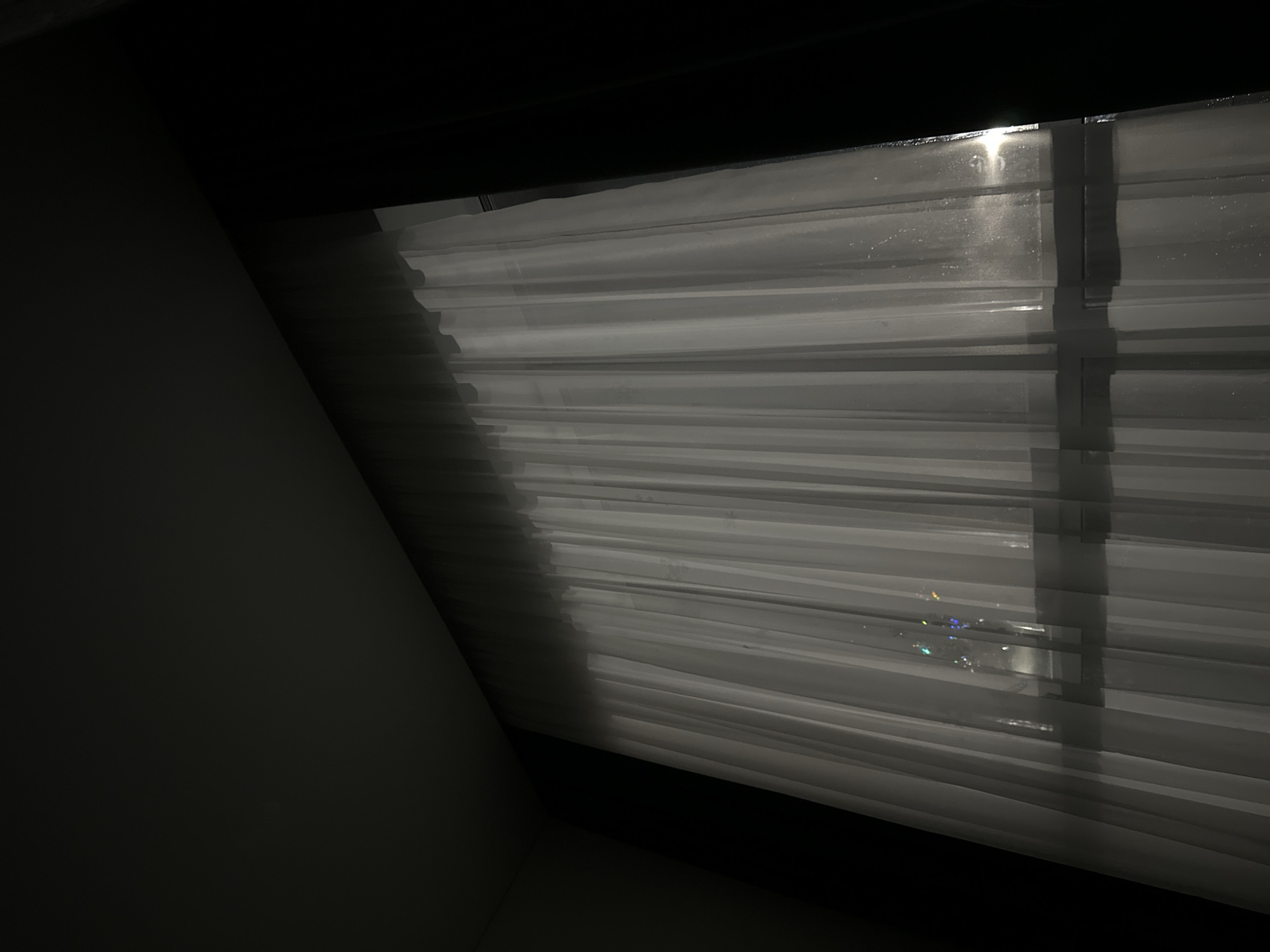 The image depicts a partially obscured view of light filtering through closed horizontal blinds. The scene is dark, with the blinds appearing white or light gray. Sunlight shines through the blinds, creating bright vertical lines and some areas of glare. A section of a dark wall is visible on the left side of the image.