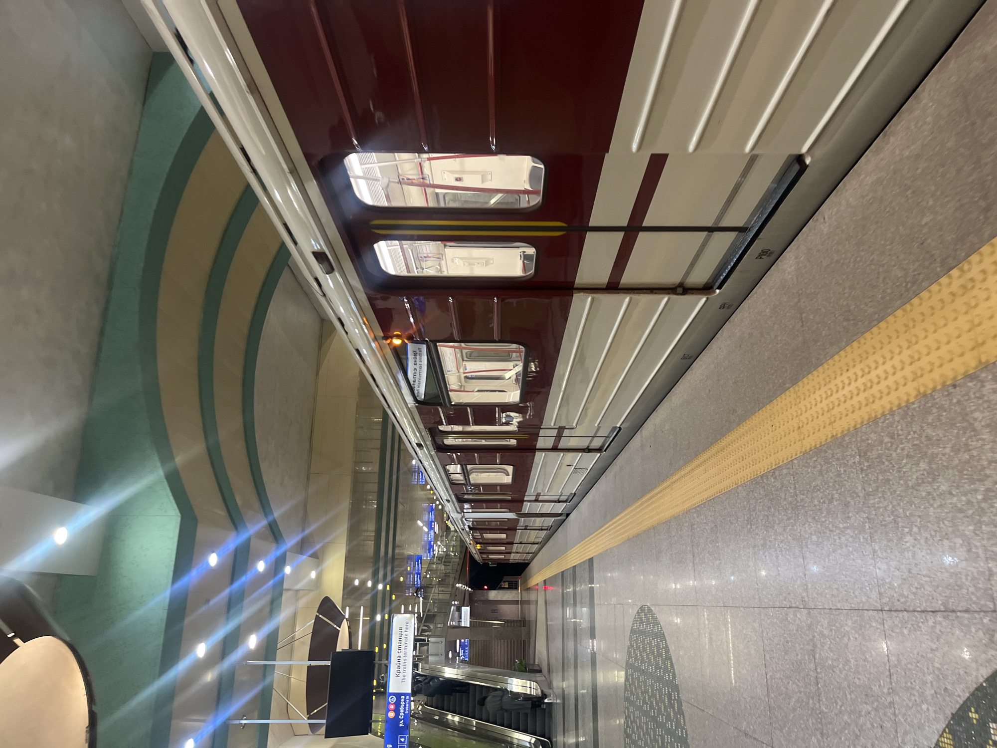 The image depicts the Sofia Metro station "Vasil Levski Stadium" in Sofia, Bulgaria. A maroon and white subway train is parked at the platform. A yellow tactile strip runs along the edge of the platform. To the left are escalator down to another level with directional signage. The wall has decorative lighting.