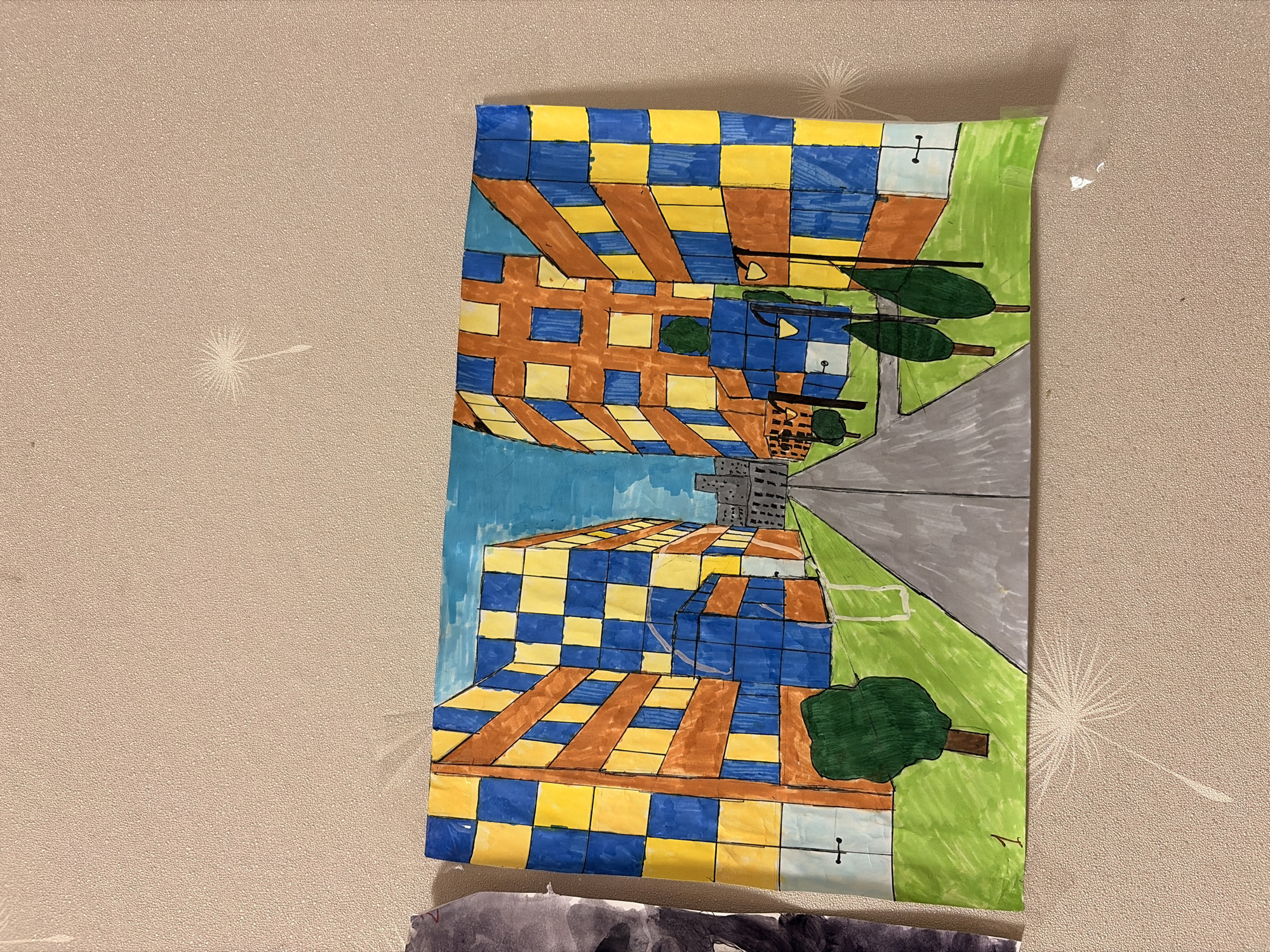The image shows a child's drawing of a city street with buildings. The buildings are colorful, with alternating blue and yellow squares on their facades. There are trees and lampposts along the street, and a grey road leads into the distance. The sky is blue. The drawing is taped to a textured wall with a dandelion-like pattern. The drawing is located in Leninogorsk, Russia.