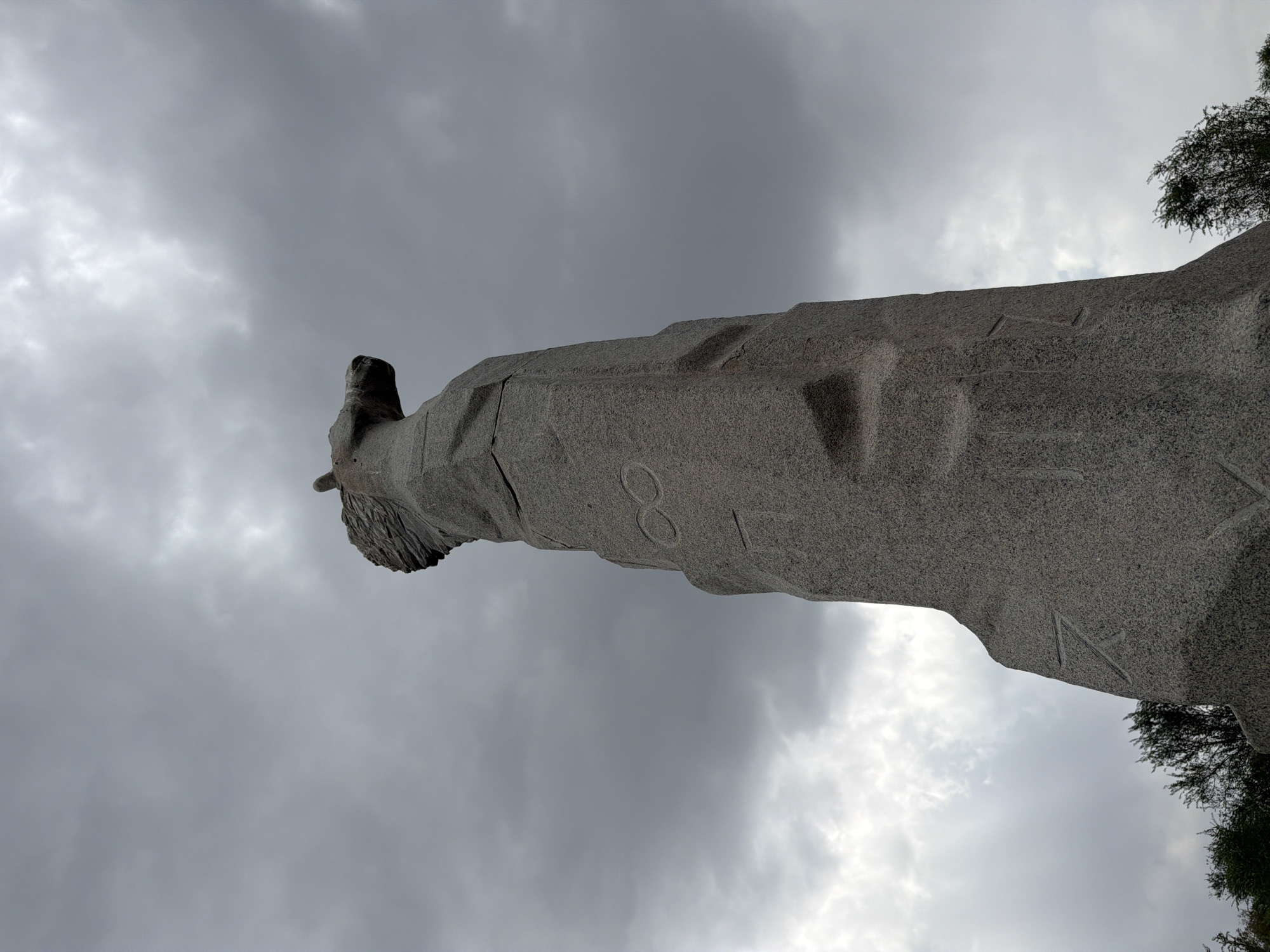 The image captures a dramatic, low-angle view of a monumental stone sculpture resembling a horse's head and neck, set against a brooding, cloudy sky. The sculpture is a rough-hewn granite structure, with its texture and sharp edges clearly visible. Symbols or runes are carved into the face of the monument. The horse's head is tilted upwards and to the right, with its mane appearing textured and windswept. The lower portion of the sculpture is massive and irregular, suggesting it is part of a larger, more abstract form. Trees are visible at the very bottom of the frame, indicating an outdoor setting. The overall atmosphere is somber and imposing, amplified by the dark clouds overhead. This sculpture is known as the "Horseman" monument and is a prominent landmark in the city of Almaty, Kazakhstan, located on Kok Tobe hill.