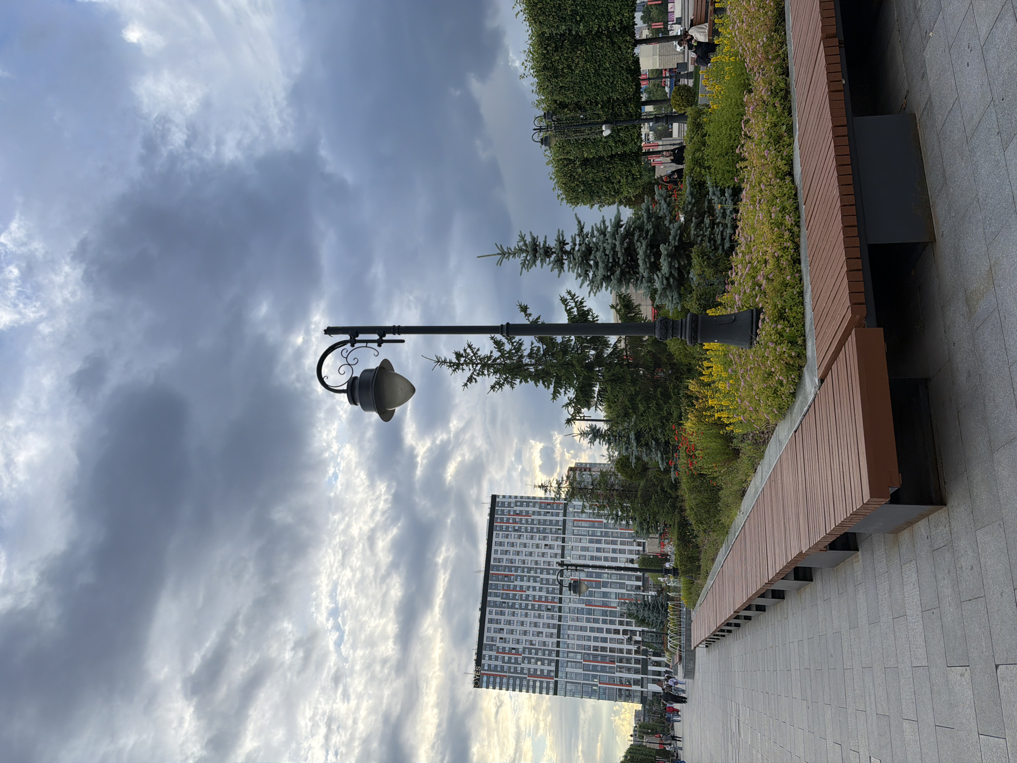 The image depicts a city park or promenade on an overcast day. In the foreground, a long, wooden slatted bench runs along the right side of a grey stone paved walkway. Beyond the bench, a meticulously landscaped garden features various evergreen trees, shrubs, and flowering plants in shades of green, yellow, and pink. A tall, black lamppost with a decorative curved arm stands prominently in the garden.
In the middle ground, a modern, multi-story building with a white facade and red and dark grey accents dominates the left side of the frame. The building appears to be an apartment complex or hotel, with numerous windows visible. A row of fountains is situated in front of the building. Several people are walking on the paved walkway in the distance.
The background is filled with a dramatic, cloudy sky. The clouds are thick and grey, with patches of brighter sky peeking through in places, suggesting the possibility of changing weather. To the right of the building, a tall, manicured hedge forms a green wall. Further to the right, another lamppost is visible, similar in design to the one in the garden. The overall impression is one of a well-maintained urban public space under a moody sky.