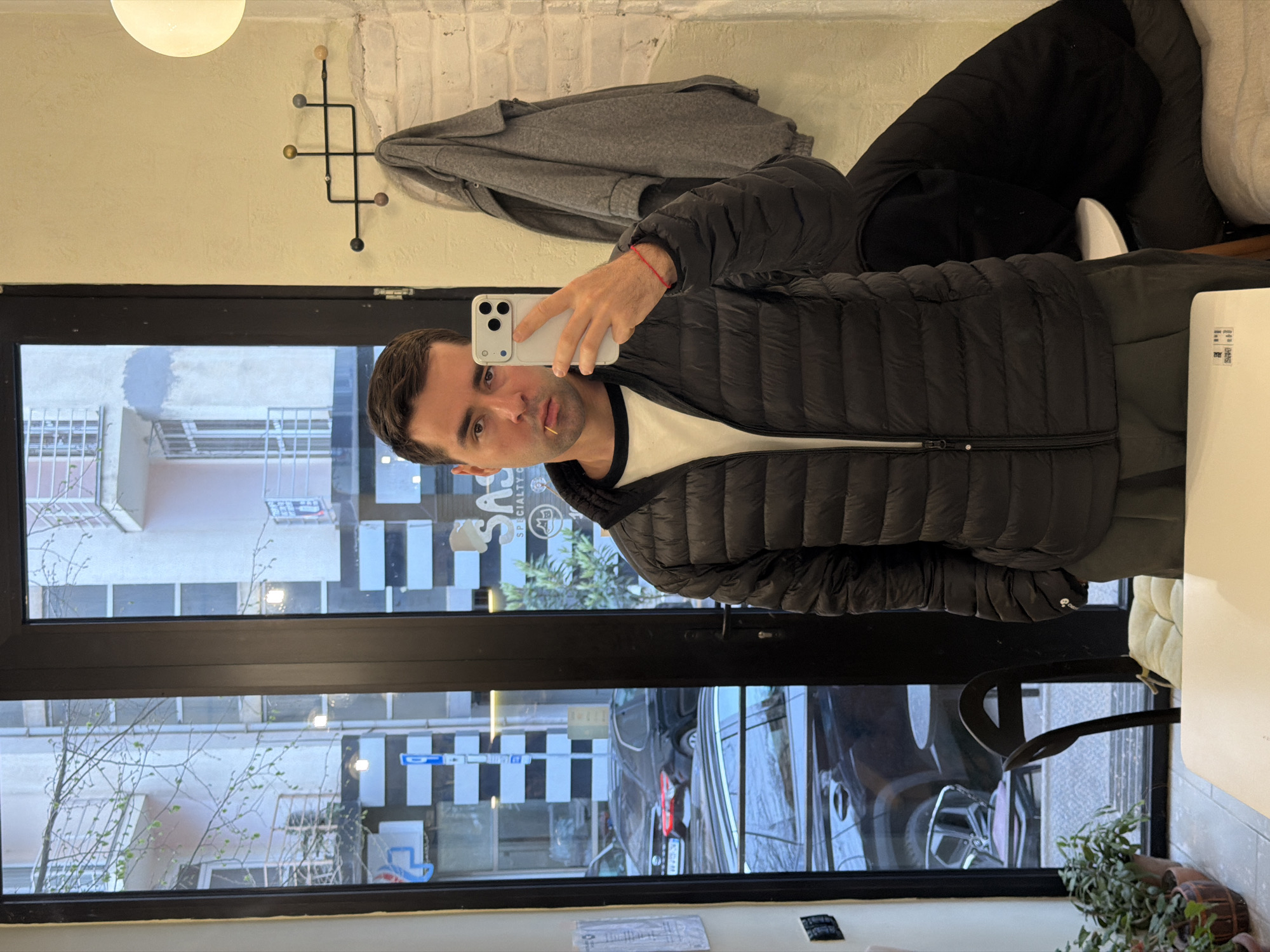 A man is taking a selfie in front of a window. The window reflects the exterior of a building, displaying a black-and-white checkered pattern. Outside, there are cars and visible street signs, potentially indicating an urban environment in Tbilisi, Georgia. The man is wearing a dark jacket over a white T-shirt and a red bracelet on his wrist. There is also a jacket hanging on a coat rack near the top of the image. A light fixture illuminates the room.