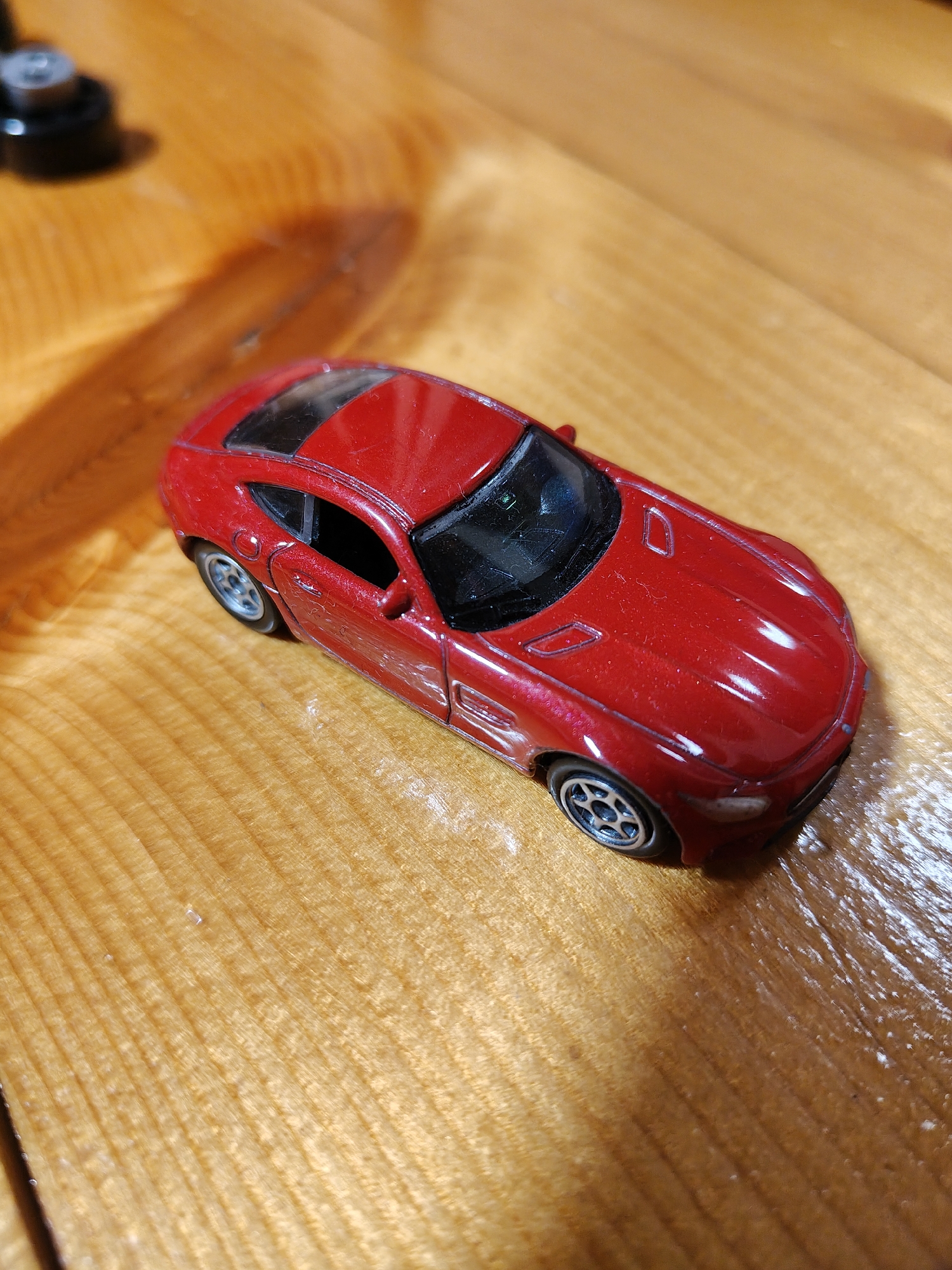 A red toy sports car is positioned on a polished wooden surface. The car has a black roof and black interior, with silver rims on its wheels. The wooden surface exhibits a visible grain pattern and is illuminated by a warm light source, casting shadows. In the upper left corner, a black object with a silver disc is partially visible. The scene is static and appears to be an indoor setting within a residential area in Фестивальное Снт, Russia.