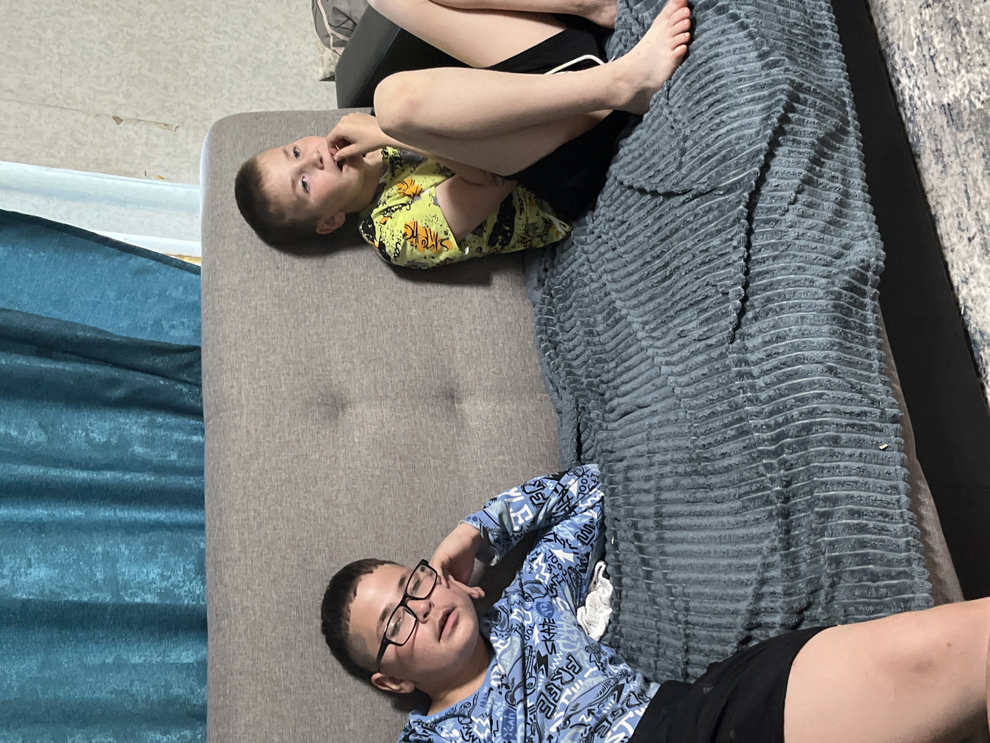 The image shows two young boys sitting on a couch in a dimly lit room. The boy on the left is wearing glasses and a blue long-sleeved shirt with a graphic print. He is leaning his head on his hand, looking to the side. The boy on the right is wearing a yellow short-sleeved t-shirt and black shorts. He is looking down and has his fingers in his mouth. The couch is covered with a dark blue textured blanket. In the background, there are blue curtains and a light-colored wall with some peeling wallpaper. The overall scene appears to be a casual indoor setting, likely a living room. The location is identified as Фоки, Russia, indicating an urban or semi-urban environment.