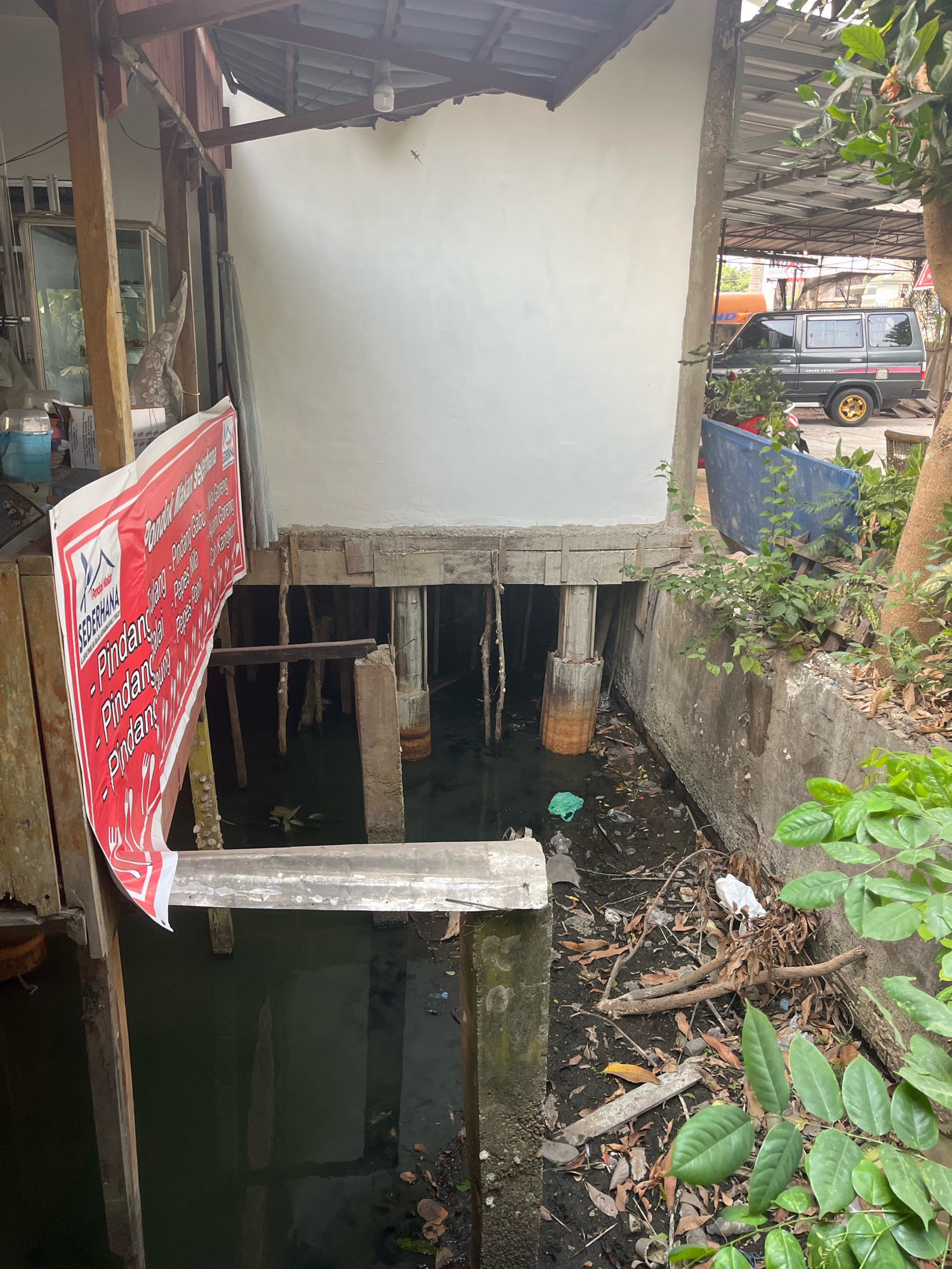 The image depicts an outdoor scene in the city of Kayu Agung, Indonesia. The foreground features a red and white banner with Indonesian text, likely advertising food, hanging from a wooden structure. To the right of the banner, a dark, stagnant body of water is visible, with debris such as leaves and plastic waste floating on the surface and along the banks. The water is contained by a concrete wall on the right side of the image. In the background, a dark-colored van is parked under a covered area. The overall environment appears to be an outdoor area adjacent to a building, possibly a residential or commercial establishment, with elements of both natural vegetation and man-made structures. The lighting suggests it is daytime.