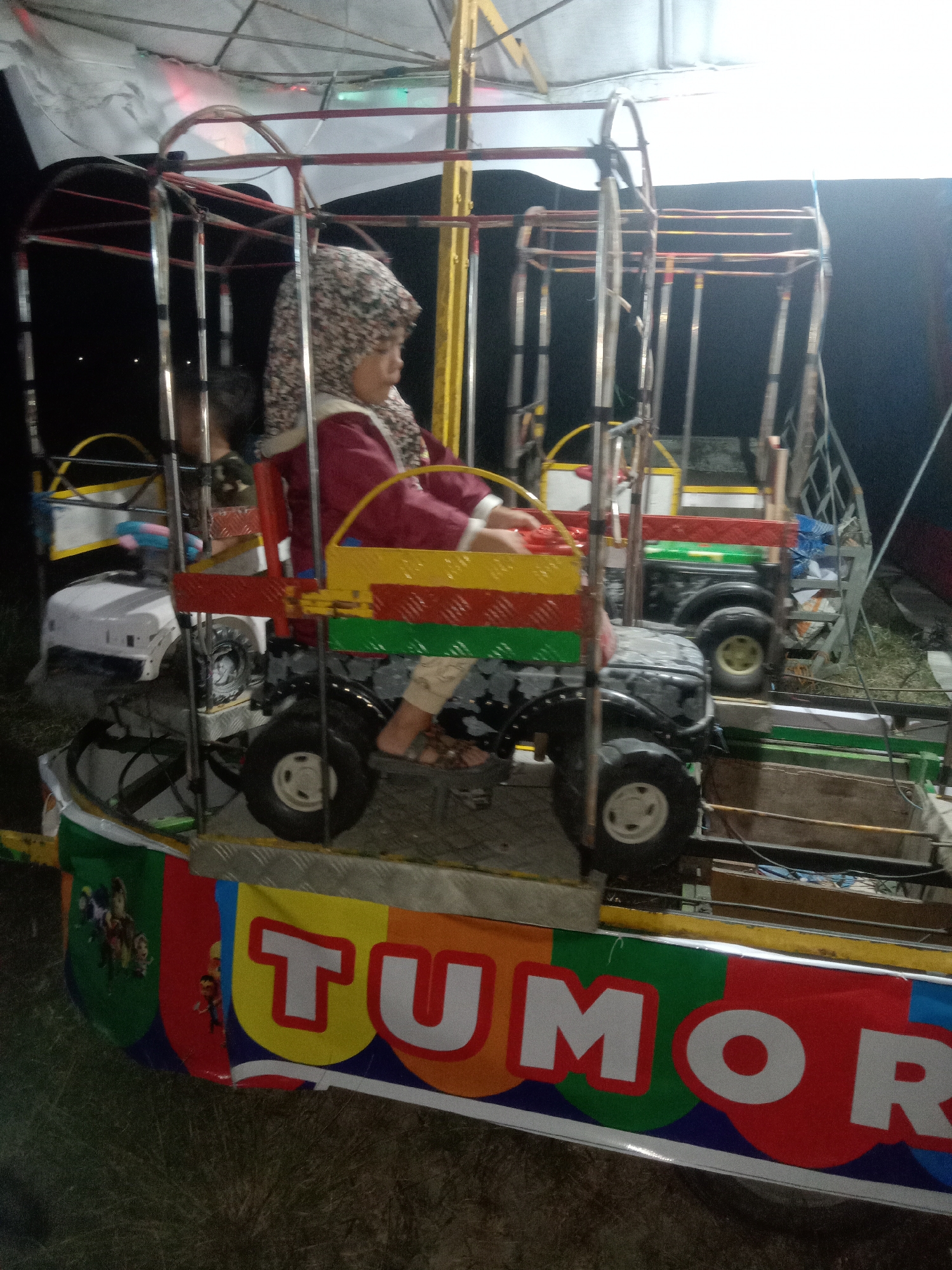 The image depicts an outdoor amusement ride at night, possibly in the city of Jumput, Indonesia. The ride features several small, stylized jeep-like vehicles on a circular track, some of which are decorated with bright colors and cartoon characters. A young child wearing a headscarf and a maroon jacket is seated in one of the vehicles, appearing to be steering. The ride is illuminated by artificial lights, casting shadows on the surrounding grassy ground. A banner at the bottom of the ride displays the word "TUMOR" in large, colorful letters, which may be the name of the amusement or a local establishment. The overall scene suggests a local fair or carnival atmosphere.