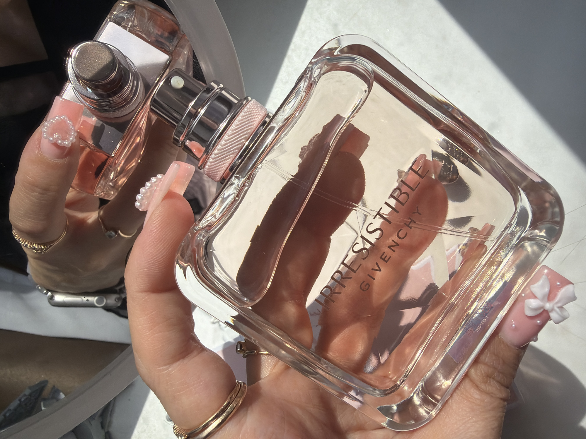 A close-up, high-angle view depicts two hands holding two perfume bottles against a light background with strong sunlight creating angular shadows. The foreground hand, with tan skin, is holding a large, clear glass bottle of "IRRESISTIBLE GIVENCHY" perfume, filled with a pale pink liquid. This hand displays multiple gold rings on the ring and pinky fingers, and elaborately manicured, square-shaped pink nails adorned with 3D white elements: a bow on the pinky, multiple pearls on the middle finger, and a heart shape made of pearls on the ring finger. The perfume bottle features a silver-toned sprayer with a textured pink band below it. The background hand, also with tan skin, holds a smaller, similar perfume bottle, and wears an Apple Watch with a silver band on the wrist, along with several gold rings on the fingers, one featuring multiple stones. The setting is indoors, with a bright, reflective surface below the objects. The location of Novosaratovka, Russia, cannot be determined from the visual information provided.