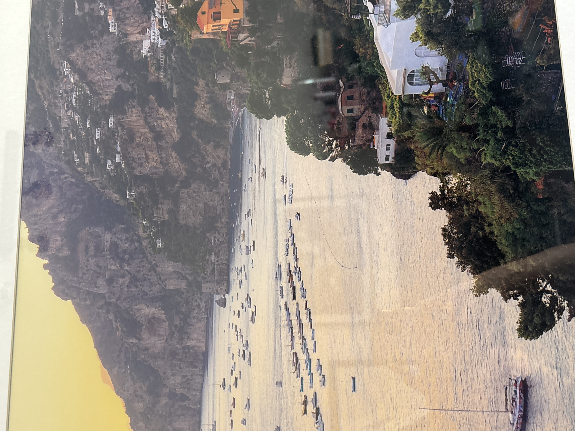 The image depicts a picturesque coastal scene with a town built into a steep, rocky mountainside overlooking a body of water filled with numerous boats. The architecture features white buildings with orange and yellow accents, nestled among lush green vegetation. The sun appears to be setting or rising, casting a warm, golden light on the water and the landscape. Several boats, ranging from small dinghies to larger yachts, are anchored or cruising in the bay. The mountainous terrain is dotted with houses and structures, giving the impression of a Mediterranean village. The foreground shows a cliff edge with more buildings and greenery, including lounge chairs and umbrellas, suggesting a resort or residential area. The water is calm and reflects the sky and surrounding scenery. There are no people prominently visible in the image. The scene is clearly not in Dubai, United Arab Emirates, but rather resembles a European coastal town, possibly in Italy.
